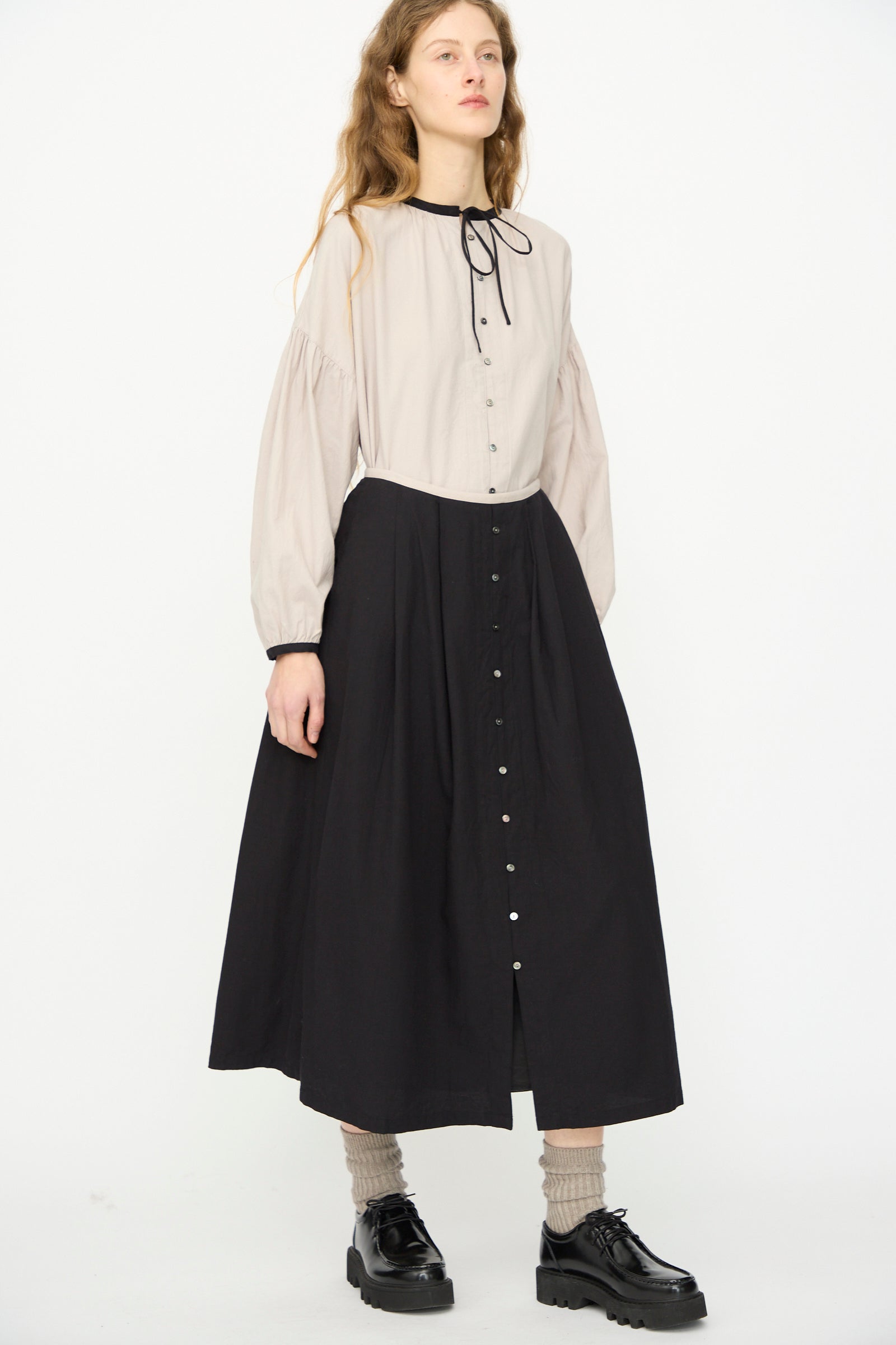 A person stands against a plain white background, wearing a long-sleeved light blouse with a black bow tie, paired with the Ichi Antiquités Cotton Cashmere Viyella Skirt in Black, along with socks and black shoes.