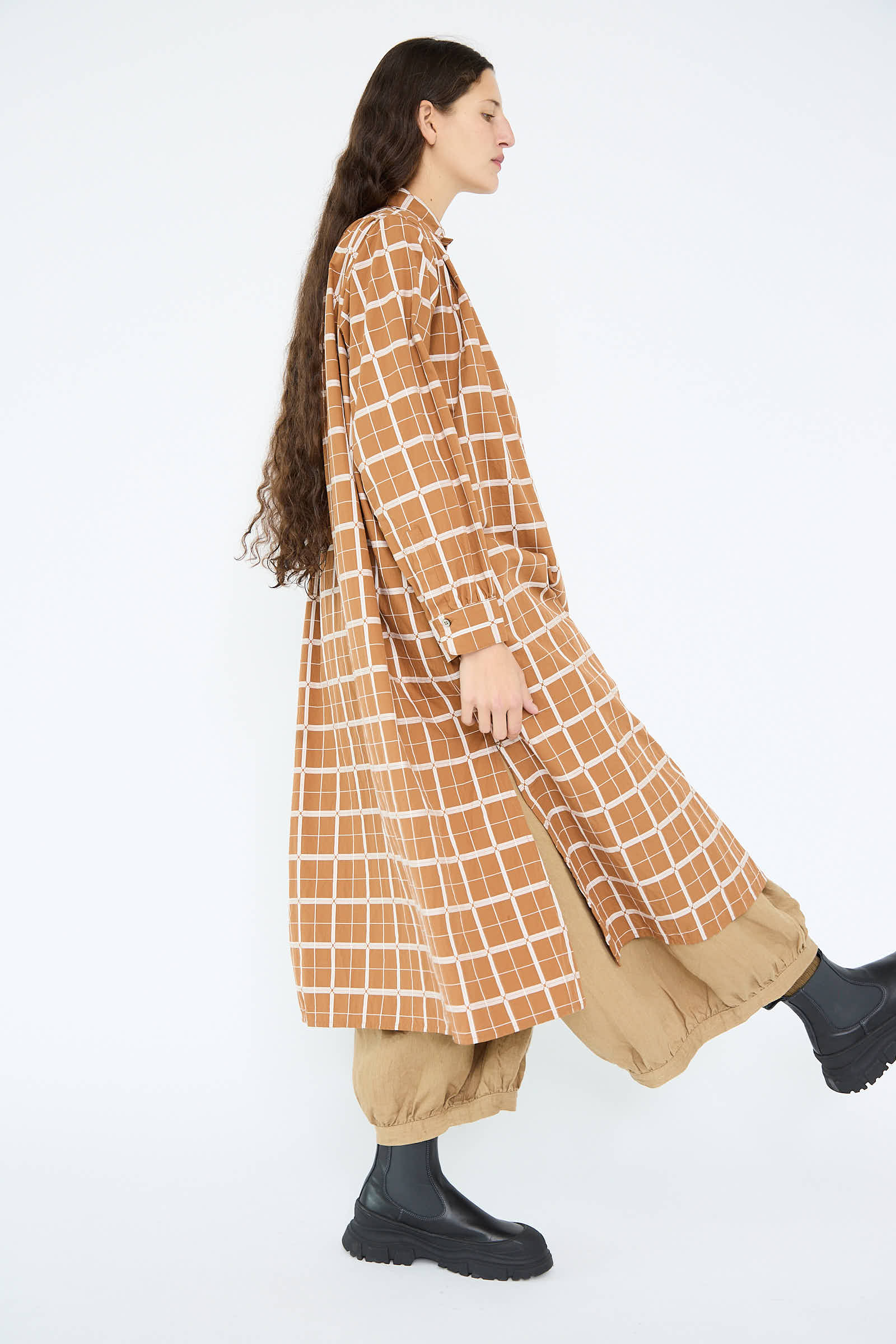 A person with long hair strides confidently in a brown Cotton Dobby Check Dress by Ichi Antiquités, paired with baggy pants and black boots.