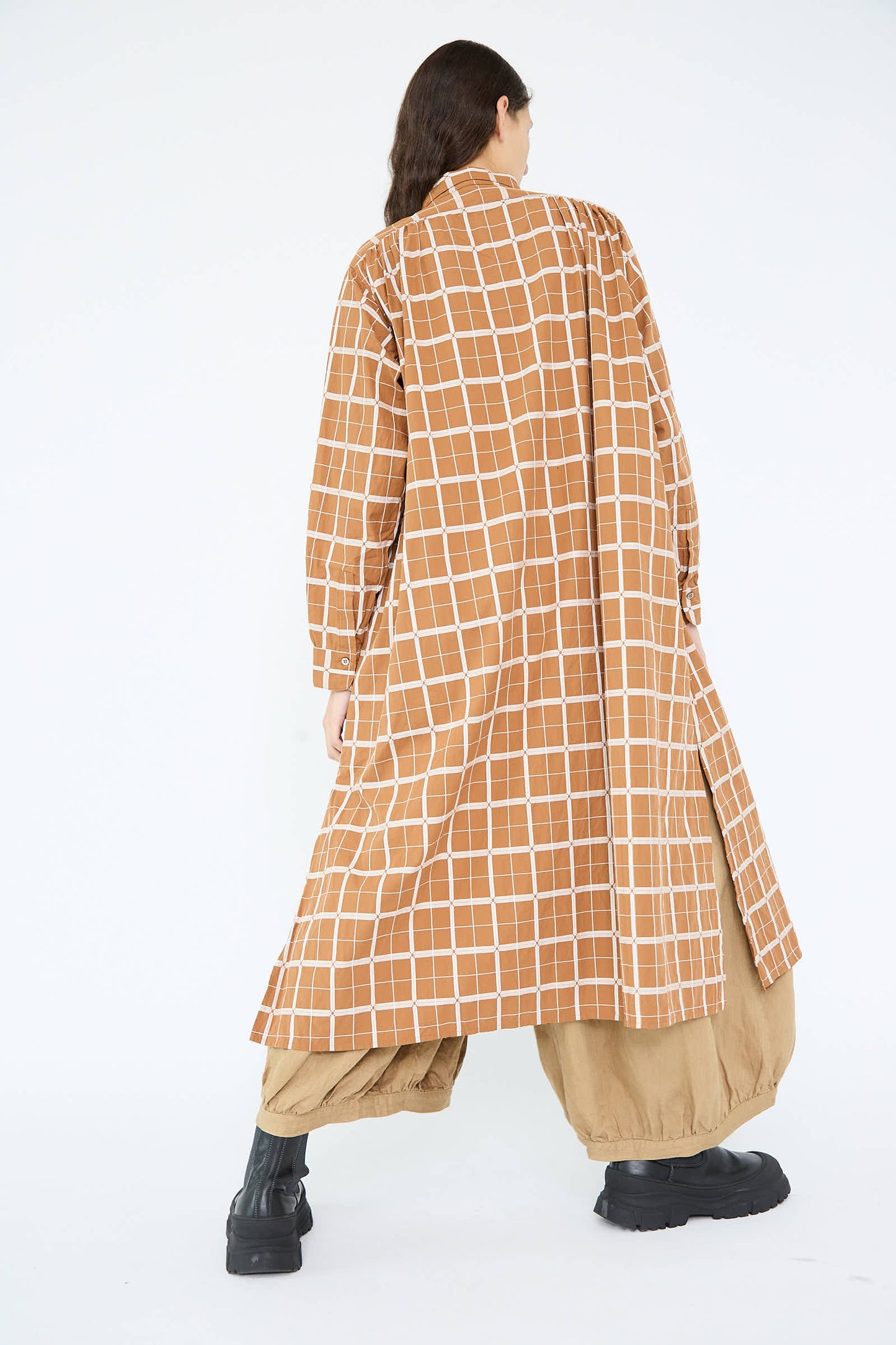 The person, wearing the Cotton Dobby Check Dress in Brown by Ichi Antiquités with wide sleeves, is paired with beige pants and black boots, facing away from the camera.