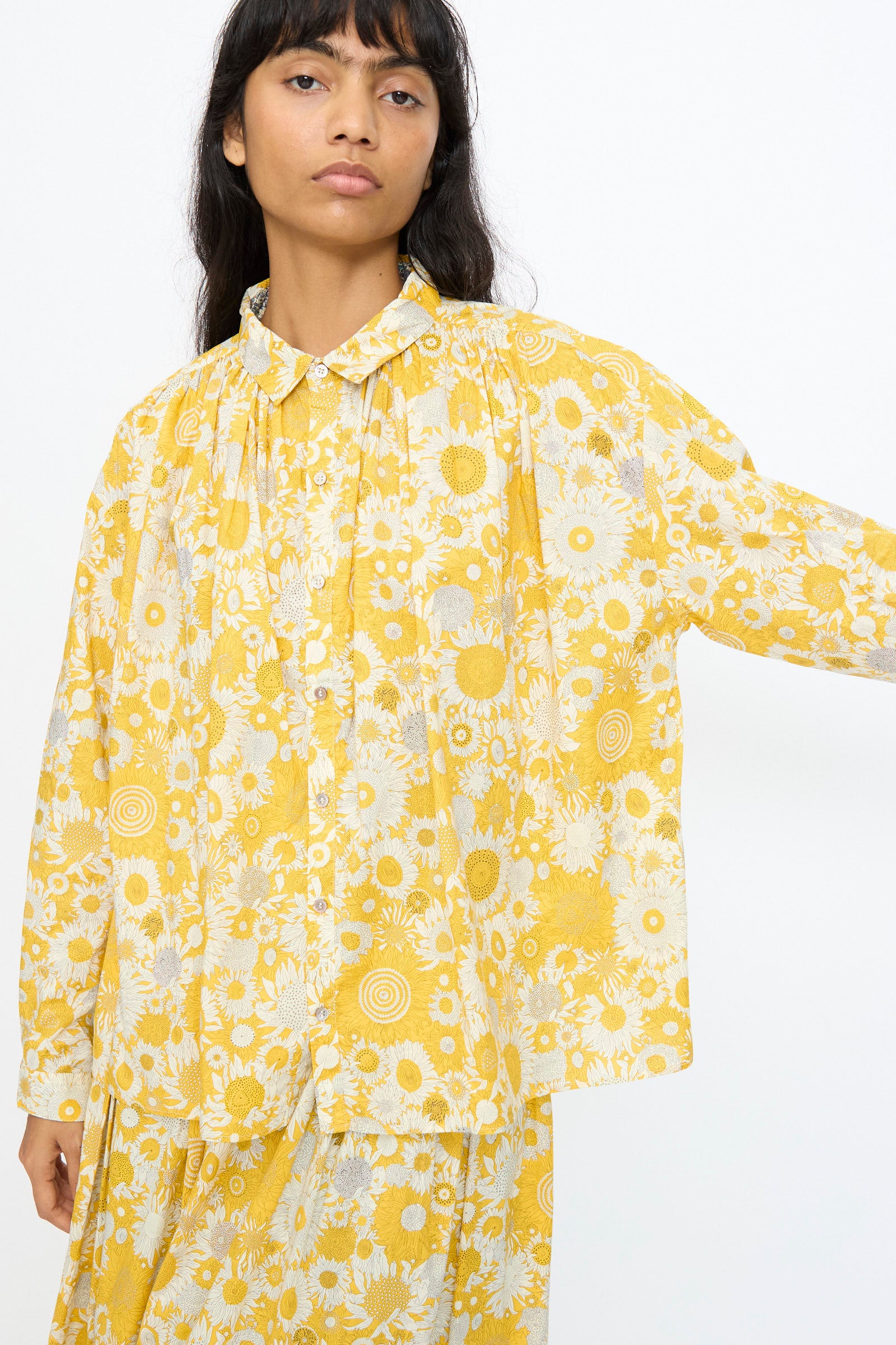 A person wears the Ichi Antiquités Cotton Liberty Shirt in Yellow, styled in a loose fit with long sleeves and a floral print, paired with a matching skirt, standing against a plain white background.