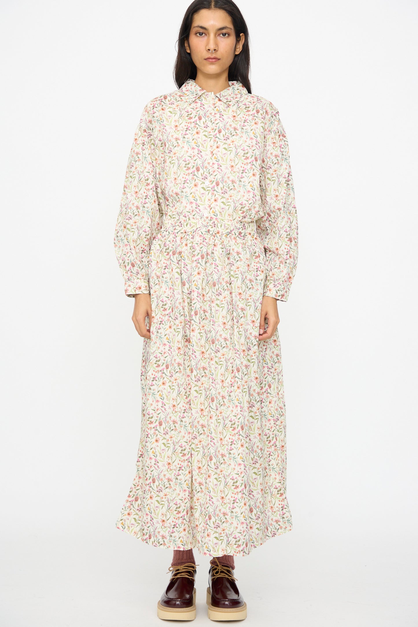 A person stands against a plain white background wearing the Ichi Antiquités Cotton Liberty Skirt in Flower Print paired with a long-sleeved top and burgundy lace-up shoes.