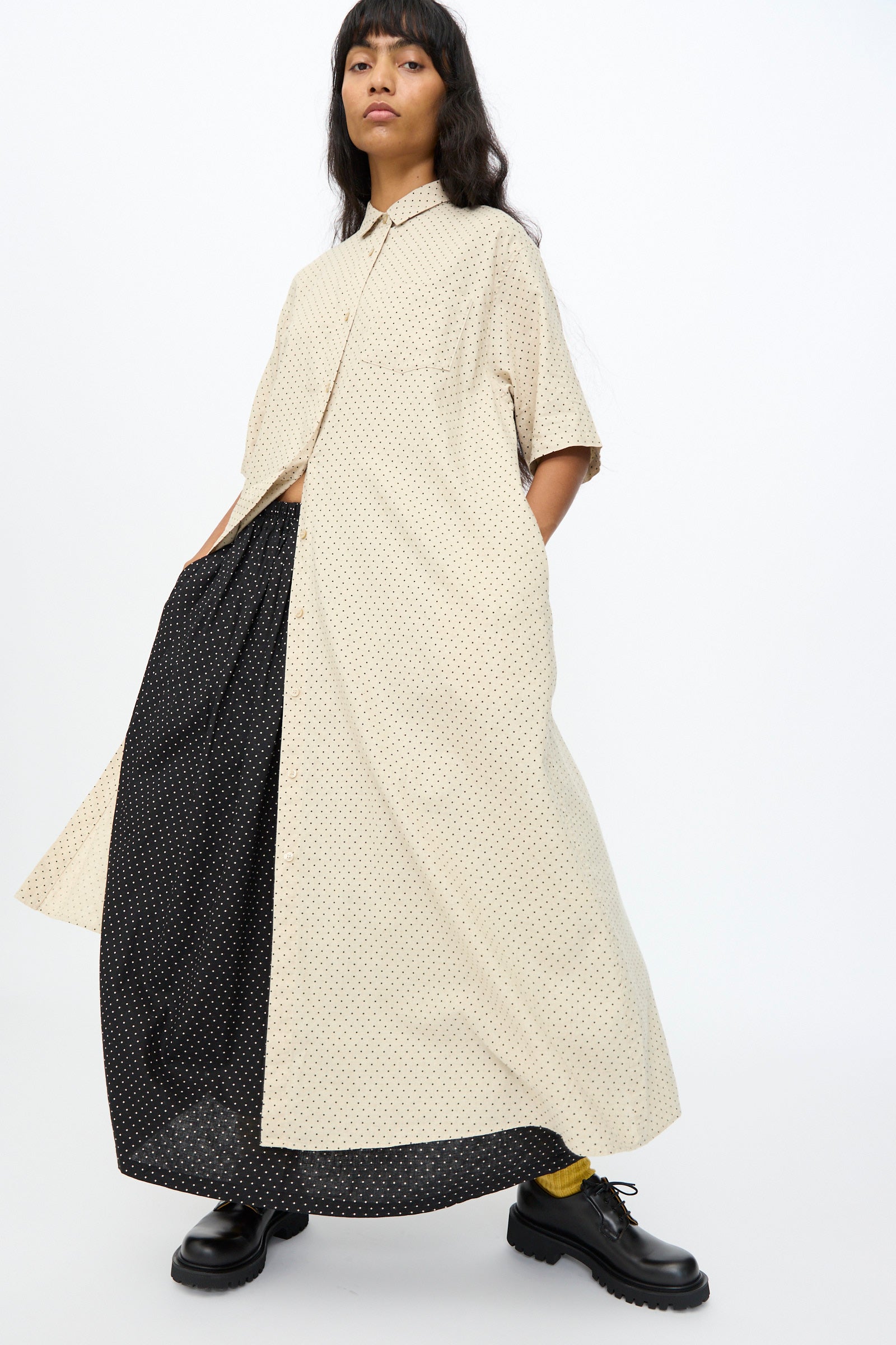 A person wears the Ichi Antiquités Cotton Linen Dot Dress in Natural, oversized and short-sleeved, layered over a black polka dot dress with black shoes and yellow socks, standing against a plain background.