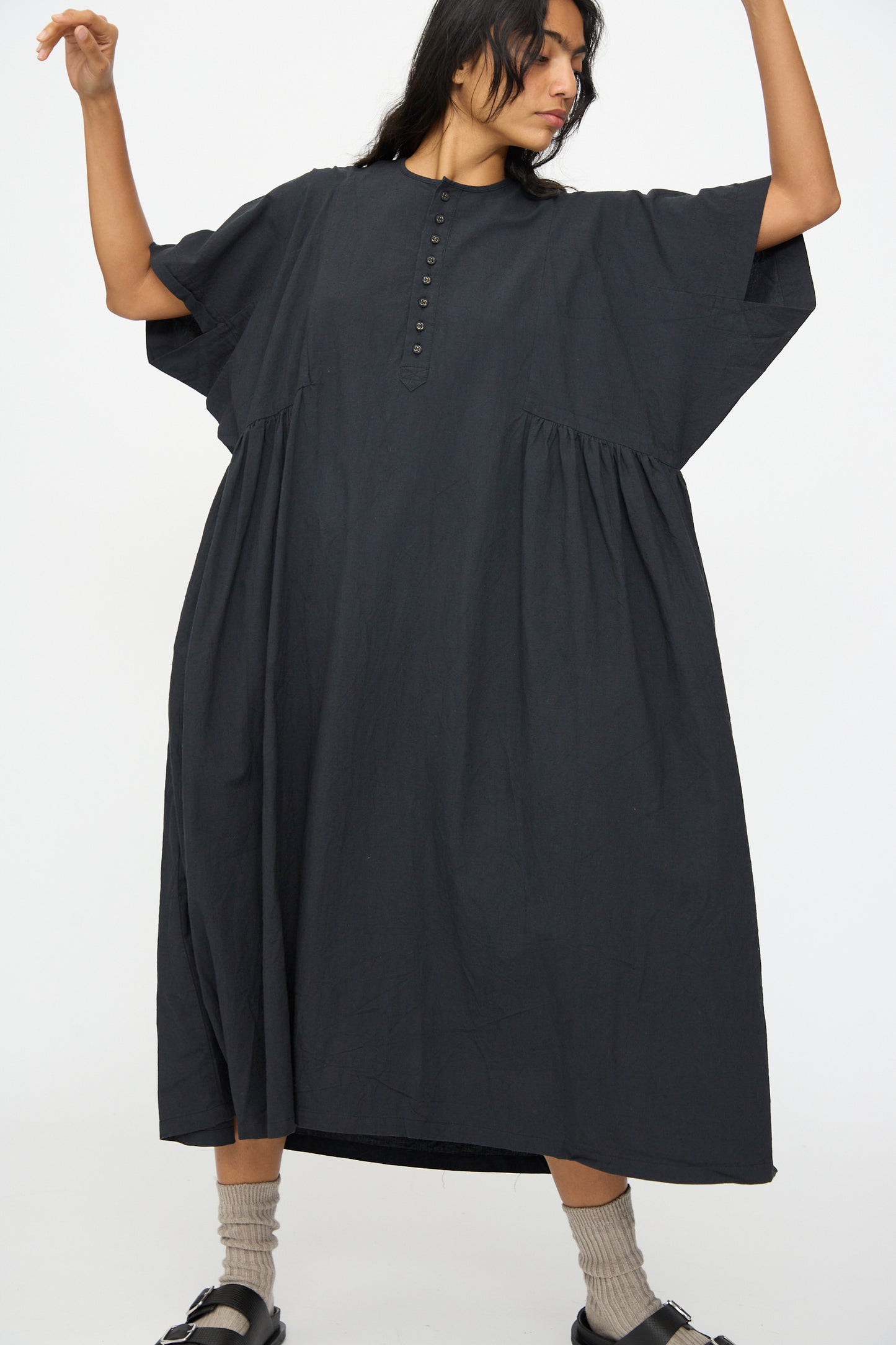 A person wears the Ichi Antiquités Cotton Linen Dress in Black, a loose-fitting short sleeve style with a buttoned front, paired with gray socks and black sandals, standing against a plain white background.