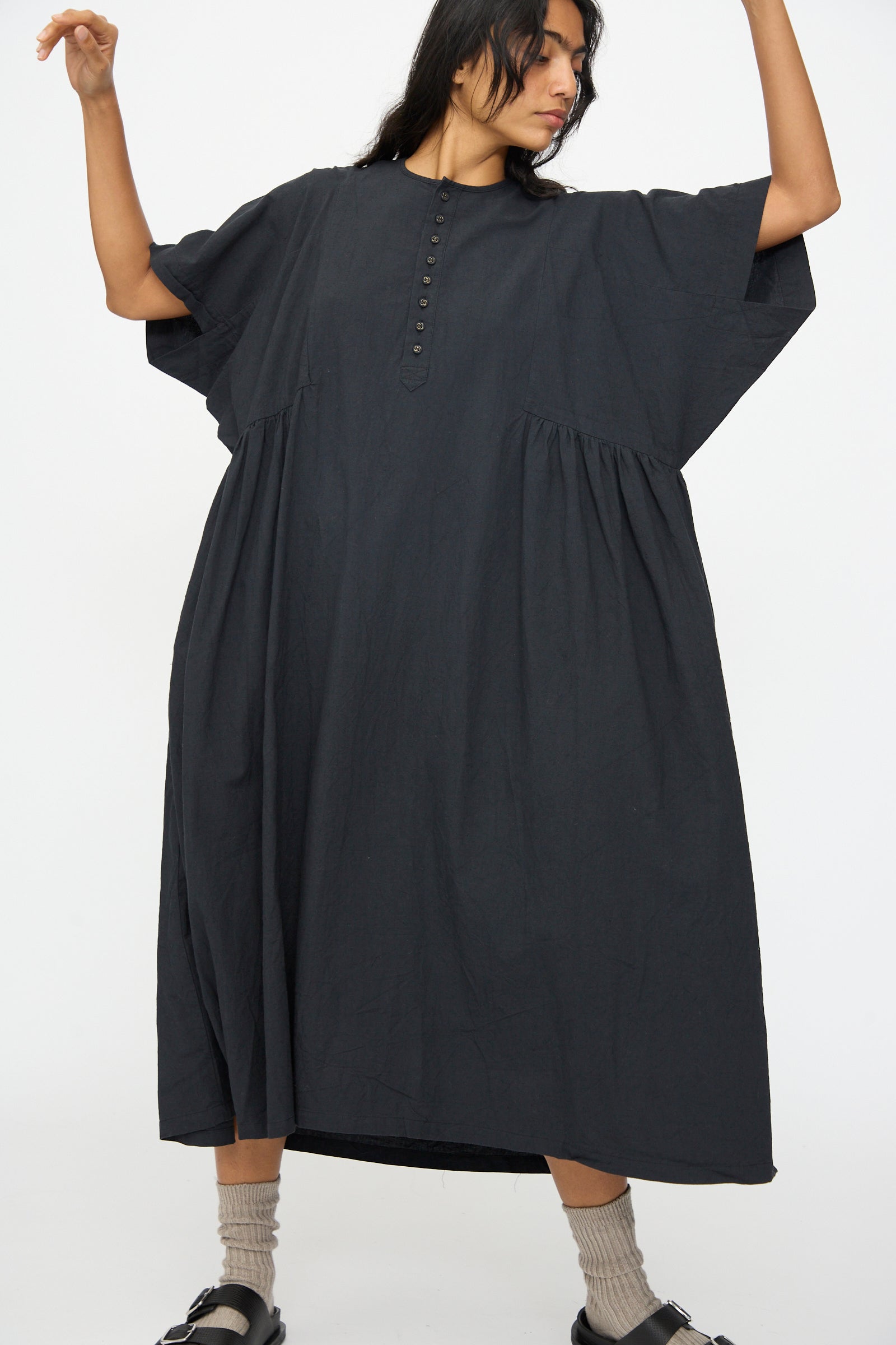 A person wears the Ichi Antiquités Cotton Linen Dress in Black, a loose-fitting short sleeve style with a buttoned front, paired with gray socks and black sandals, standing against a plain white background.