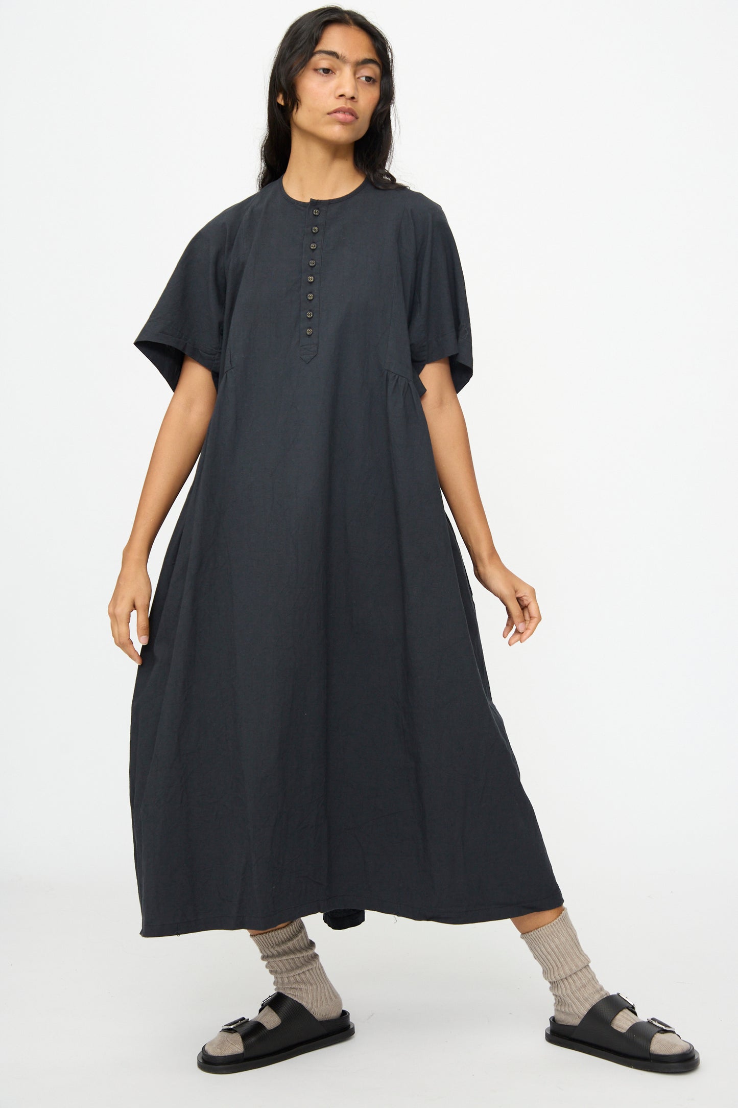 A person stands against a plain background wearing the Ichi Antiquités Cotton Linen Dress in Black, paired with grey socks and black sandals.