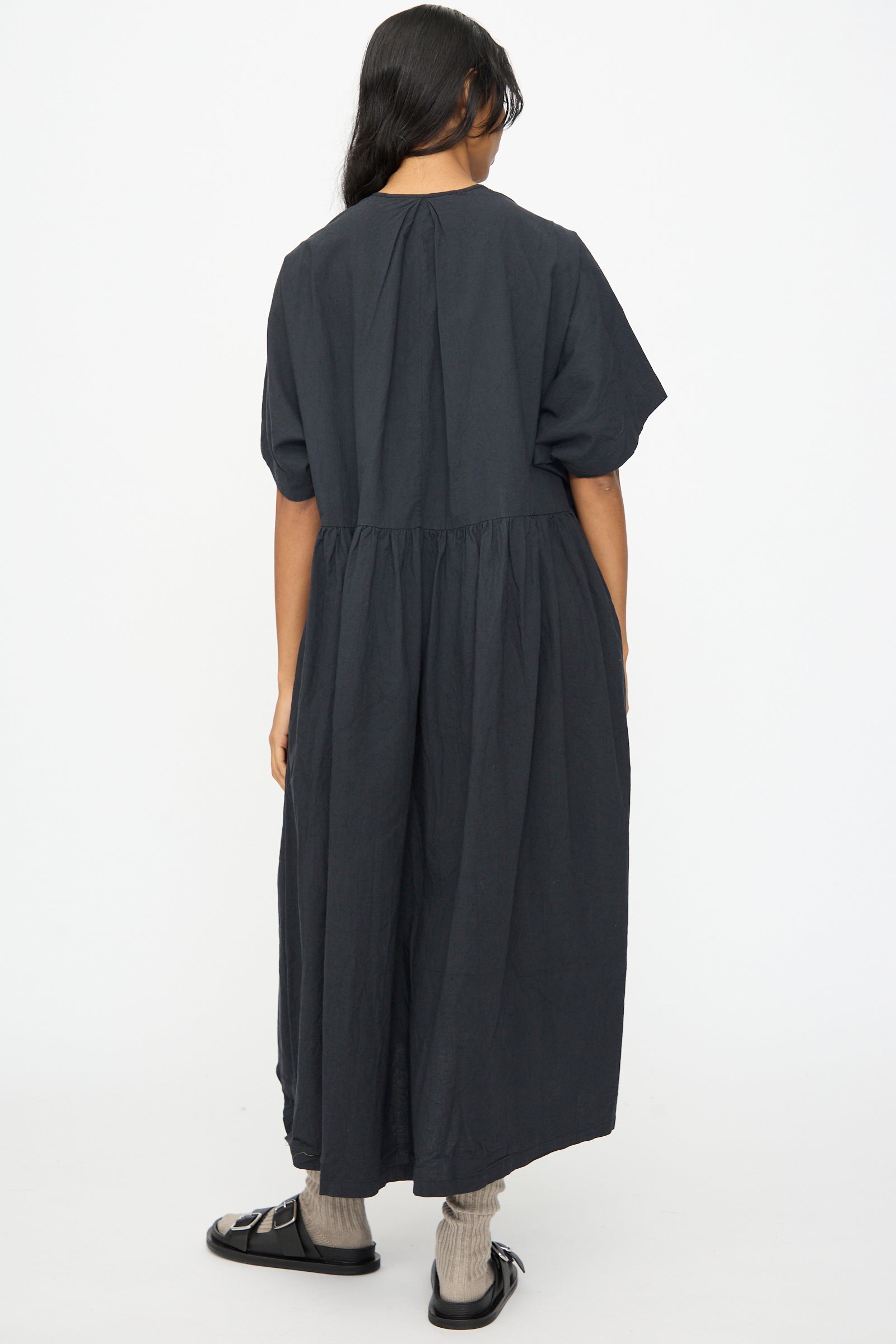 A person stands with their back to the camera, wearing the Ichi Antiquités Cotton Linen Dress in Black—an ankle-length, loose-fitting, short-sleeve style—paired with socks and black sandals against a plain white background.