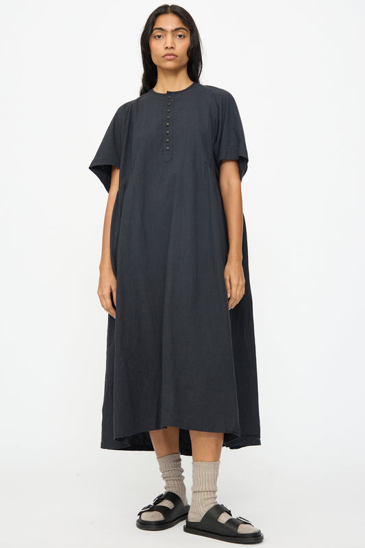 A person faces forward, wearing the Ichi Antiquités Cotton Linen Dress in Black—an oversized, short-sleeve dress with chest buttons—paired with beige socks and black sandals against a plain white background.