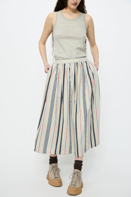 A person wearing a gray tank top and an A-line silhouette, Ichi Antiquités Cotton Linen Indigo Stripe Skirt in Multi, complemented by brown socks and beige sneakers, stands against a plain white background.