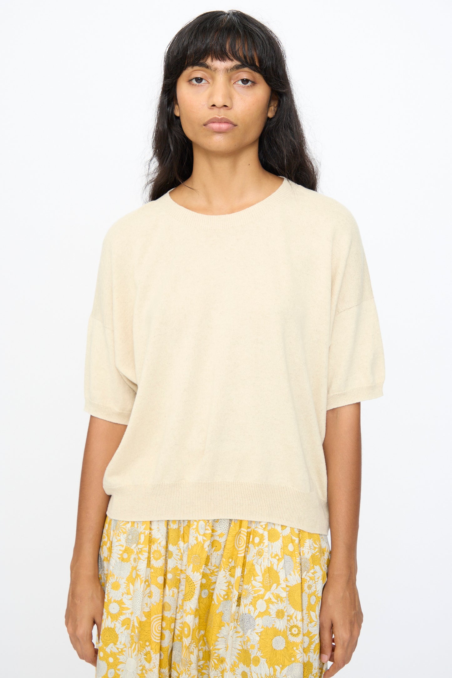 A woman with long dark hair wears the Ichi Antiquités Cotton Linen Pullover in Natural and a yellow floral skirt, standing against a plain white background.
