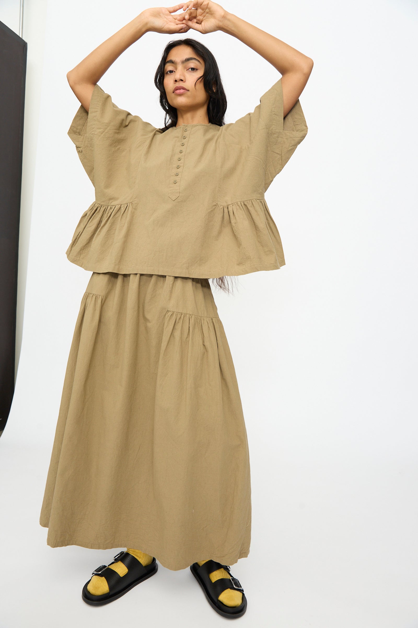 A person models the Ichi Antiquités Cotton Linen Shirt in Mocha, featuring wide short sleeves, paired with a matching long skirt, black sandals, and yellow socks against a plain white background.