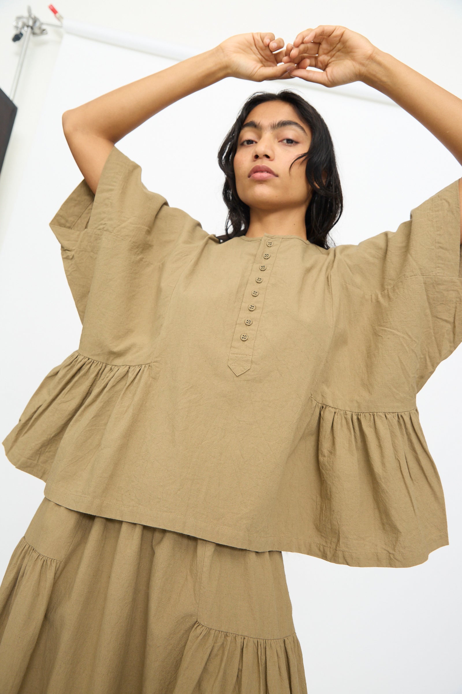 A person in the Ichi Antiquités Cotton Linen Shirt in Mocha, featuring a loose fit and wide sleeves, stands with arms raised against a plain background, styled with a matching tiered skirt.