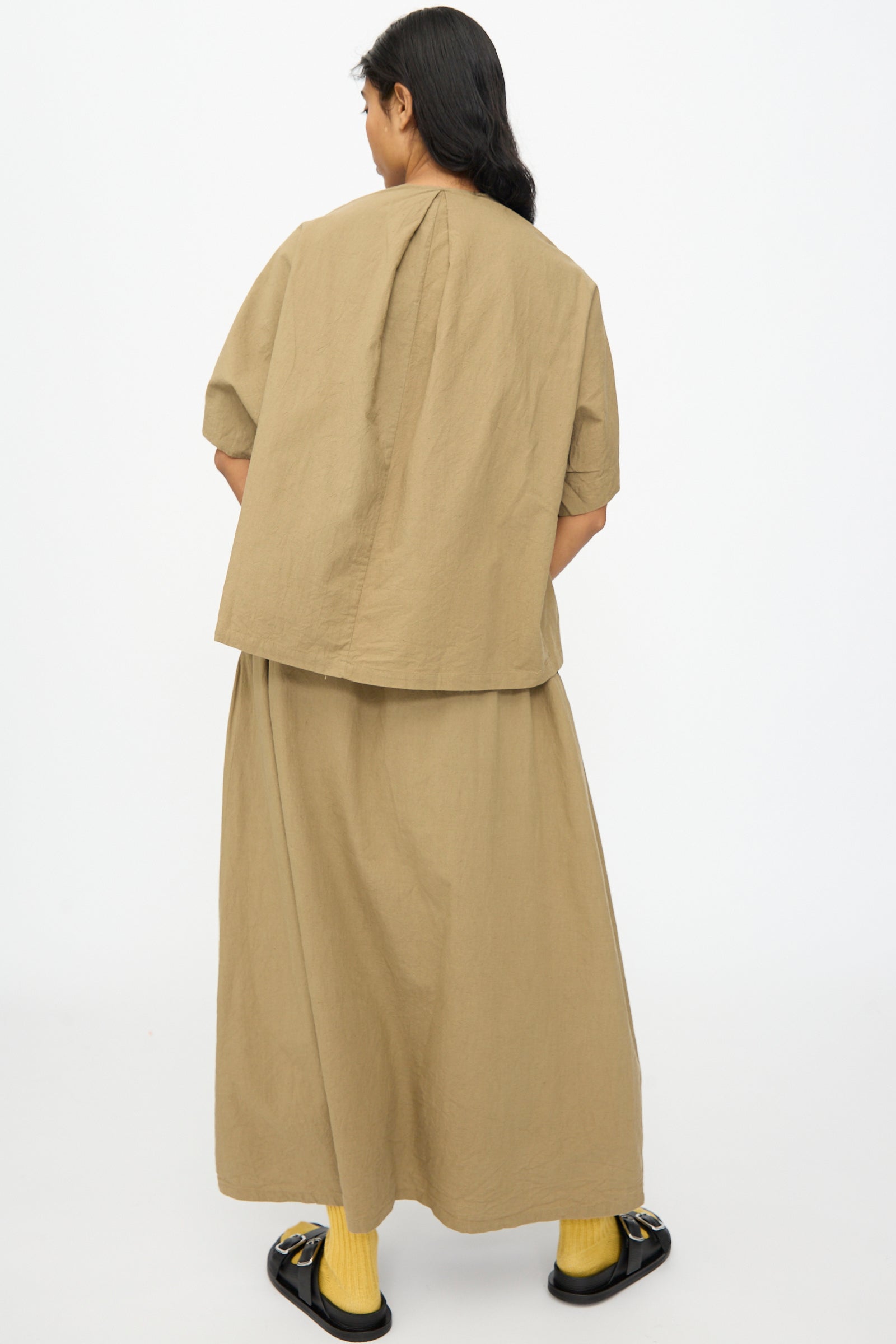 A person stands facing away against a plain white background, wearing the Ichi Antiquités Cotton Linen Shirt in Mocha with a matching long skirt, yellow socks, and black sandals.