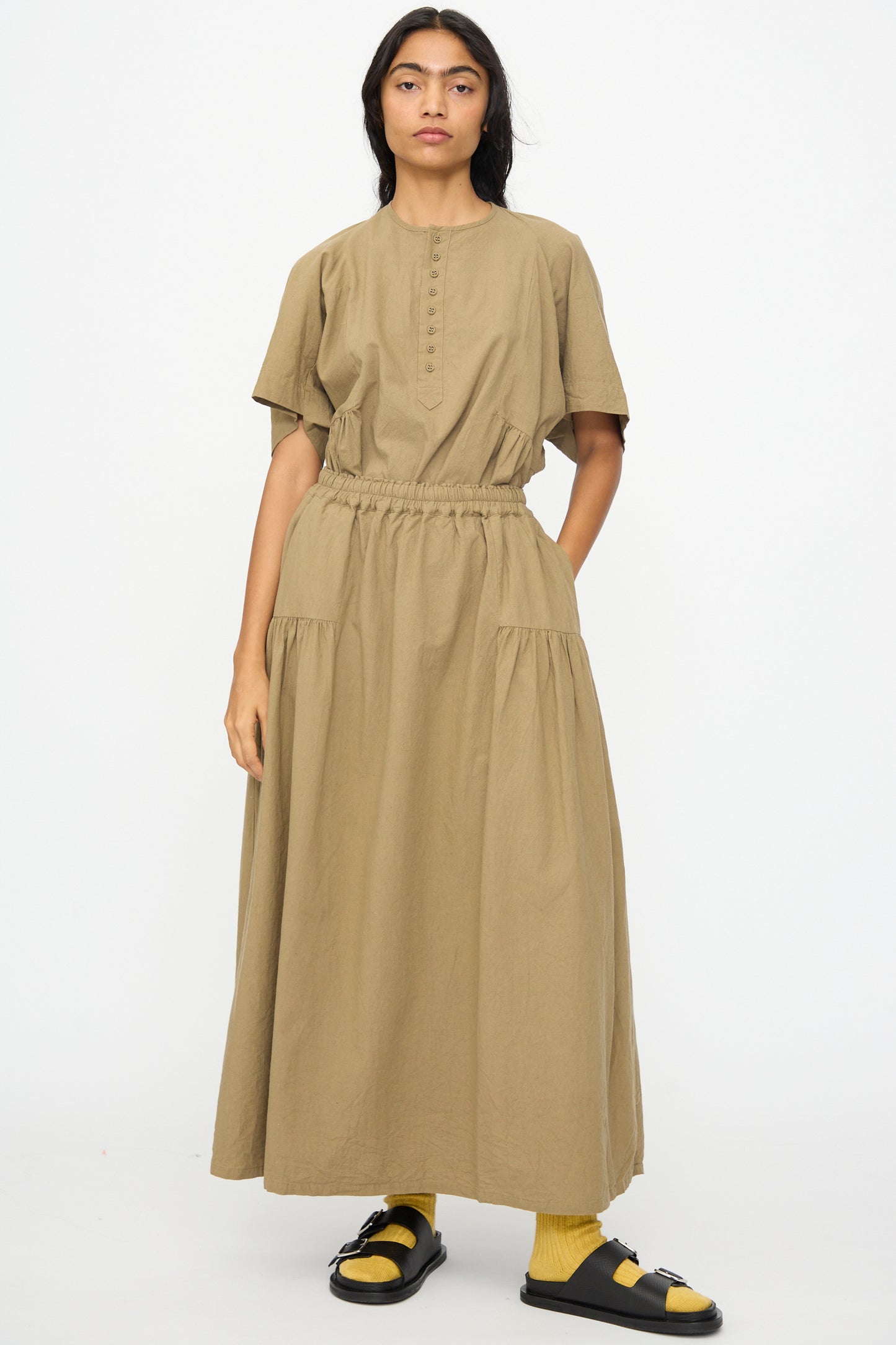 A woman faces forward against a white background, wearing the Ichi Antiquités Cotton Linen Skirt in Mocha, styled with short sleeves, yellow socks, and black sandals.