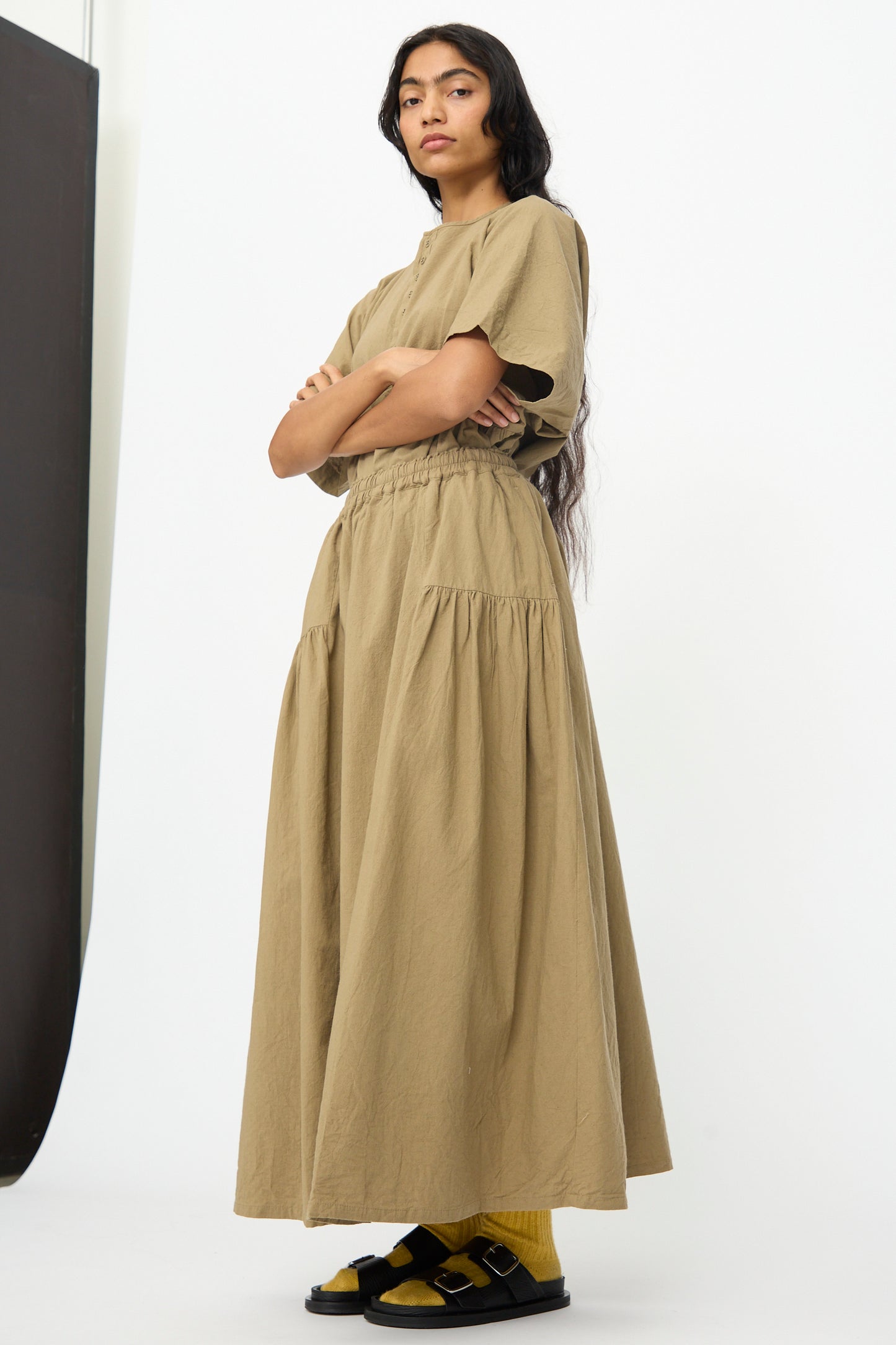 Person stands with arms crossed, wearing a loose khaki top and the Ichi Antiquités Cotton Linen Skirt in Mocha, paired with yellow socks and black sandals against a plain white background.