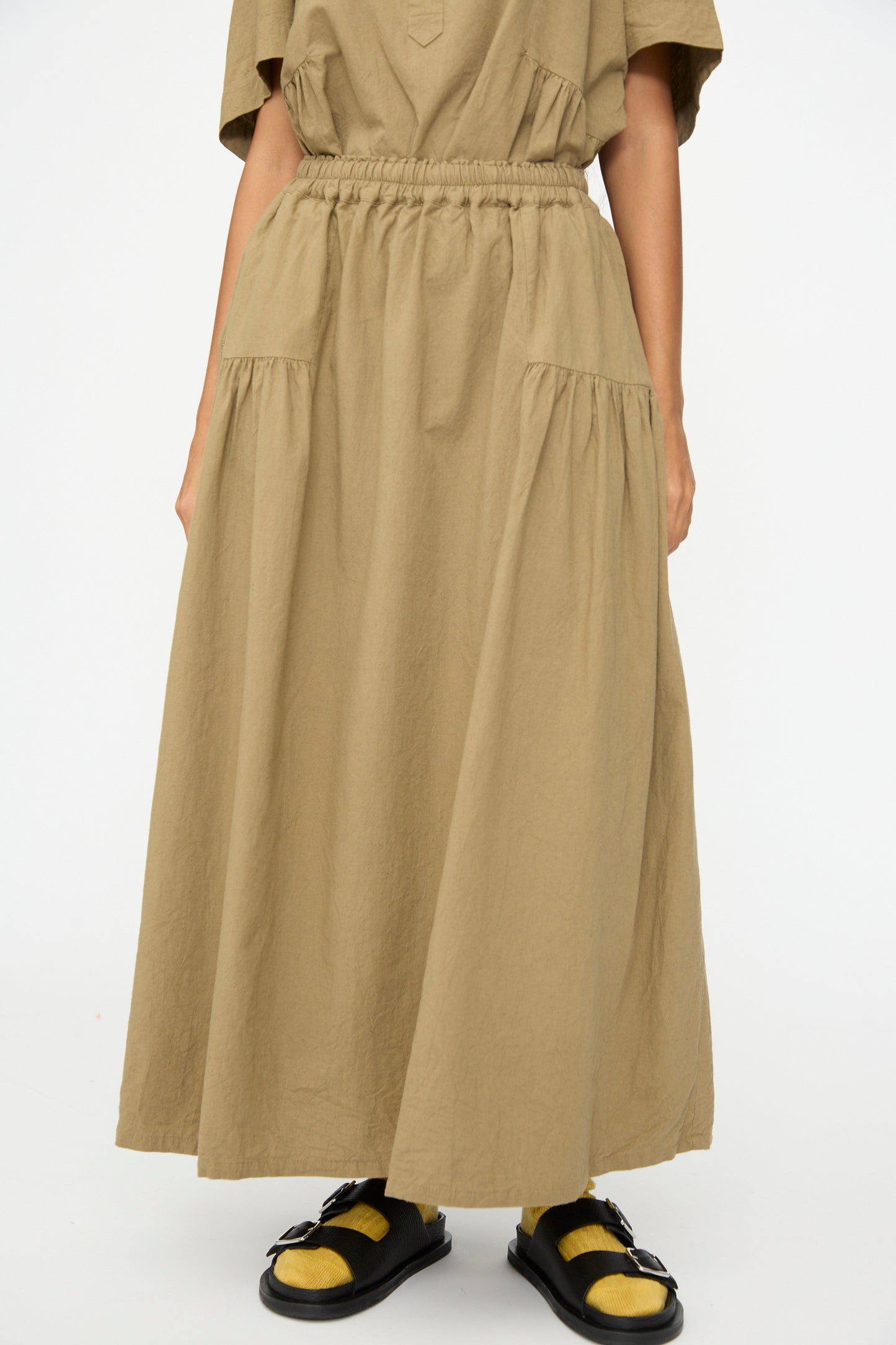 A person wears the Ichi Antiquités Cotton Linen Skirt in Mocha, a loose ankle-length design with gathered waist and side pockets, styled with black and yellow sandals. The upper body is partially visible against a plain background.
