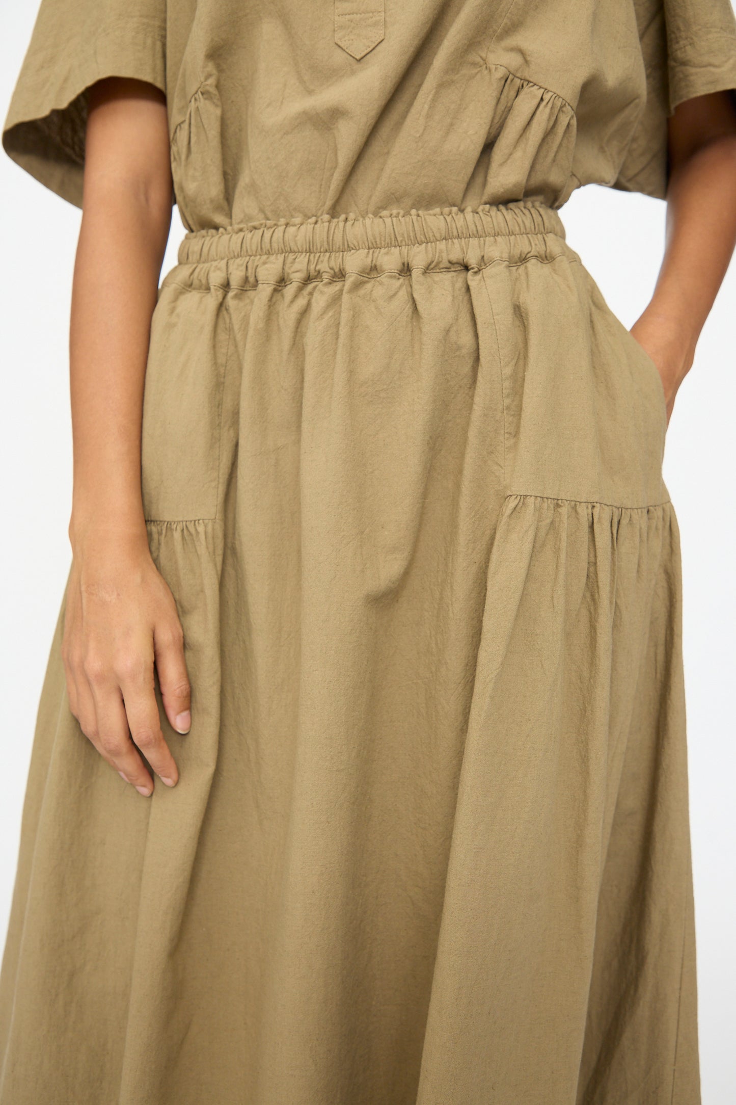 A person wears the Ichi Antiquités Cotton Linen Skirt in Mocha, a loose-fitting dress with short sleeves and an elastic waistband; one hand rests at their side while the other is tucked into a pocket.