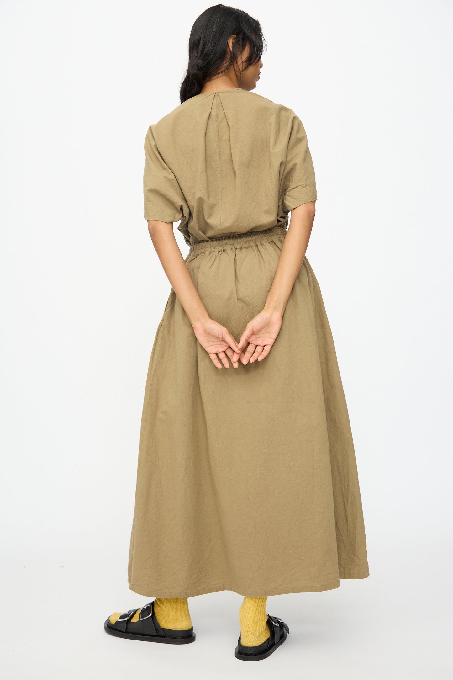 A person stands facing away, wearing the Ichi Antiquités Cotton Linen Skirt in Mocha styled as a dress, yellow socks, and black sandals, with hands clasped behind their back.