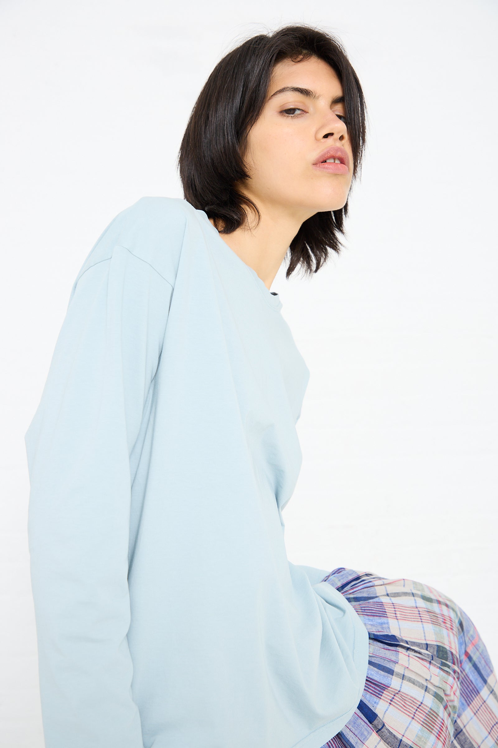 Person with short dark hair wearing an Ichi Antiquités Cotton Long Sleeve Pullover Tee in Blue and a plaid skirt, looking at the camera with a neutral expression.