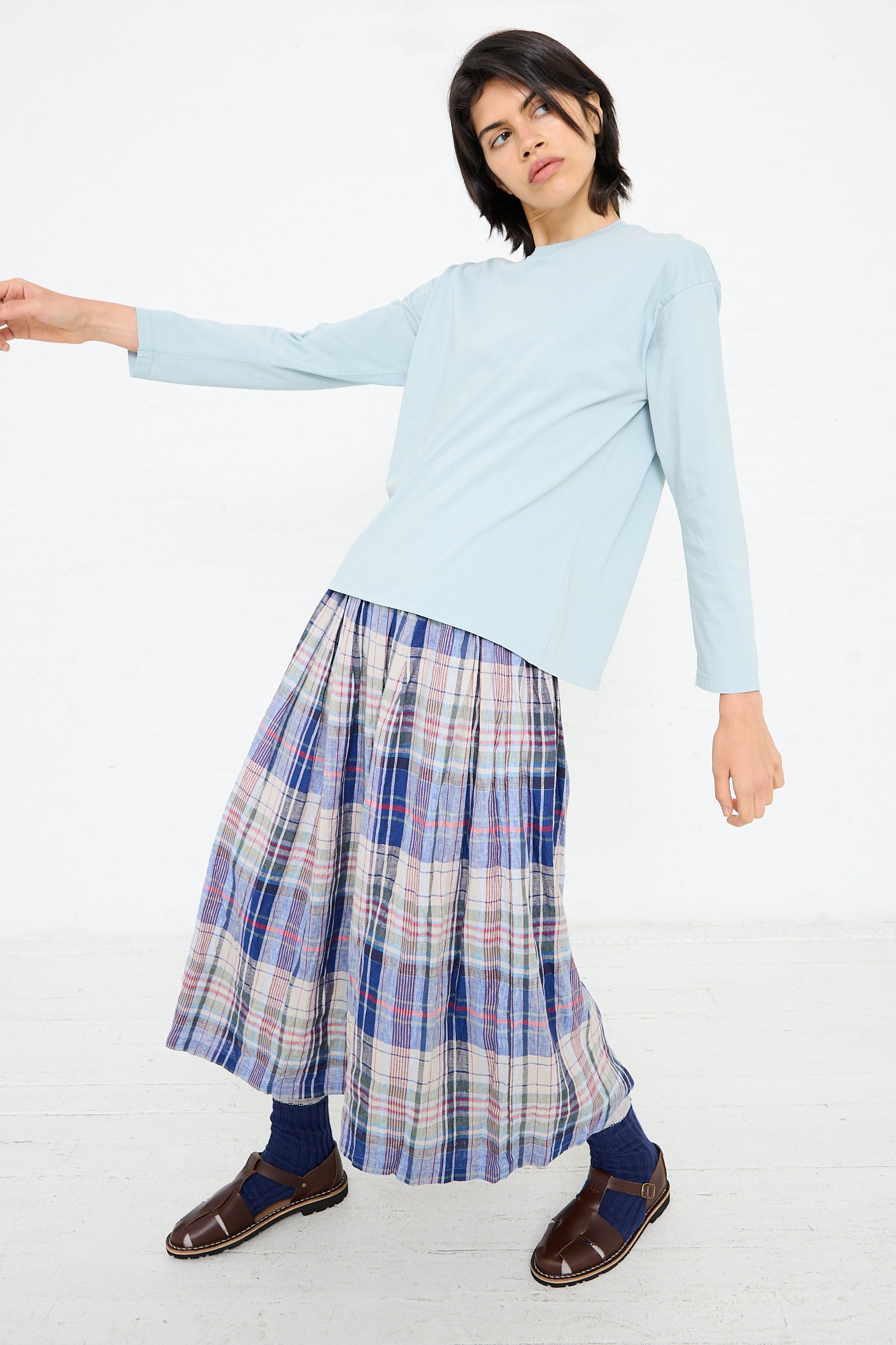 Person posing against a white background, wearing an Ichi Antiquités Cotton Long Sleeve Pullover Tee in Blue, a multicolored plaid long skirt, blue socks, and brown shoes.