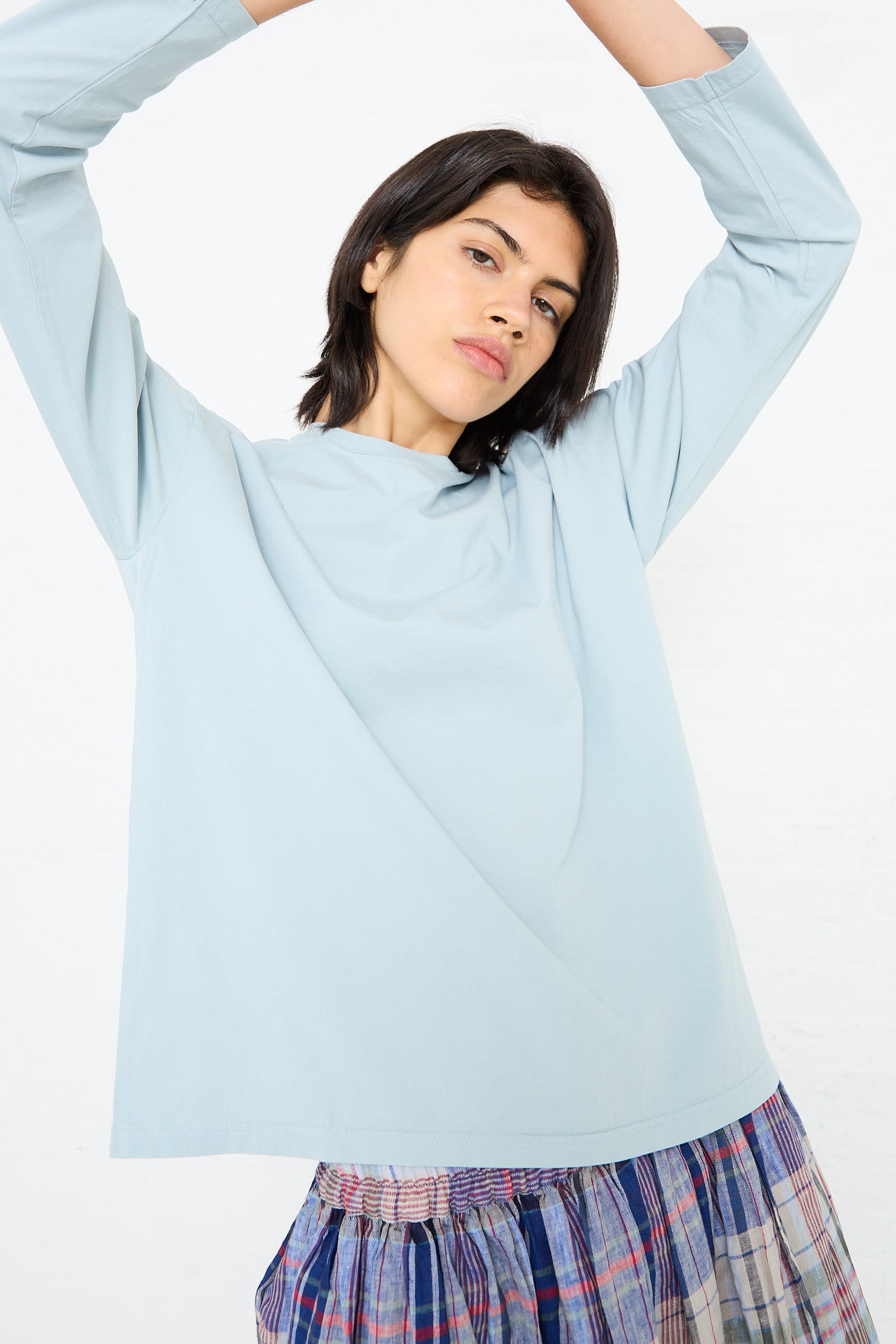 A person wearing a medium weight Cotton Long Sleeve Pullover Tee in Blue by Ichi Antiquités and a plaid skirt stretches with their arms above their head.