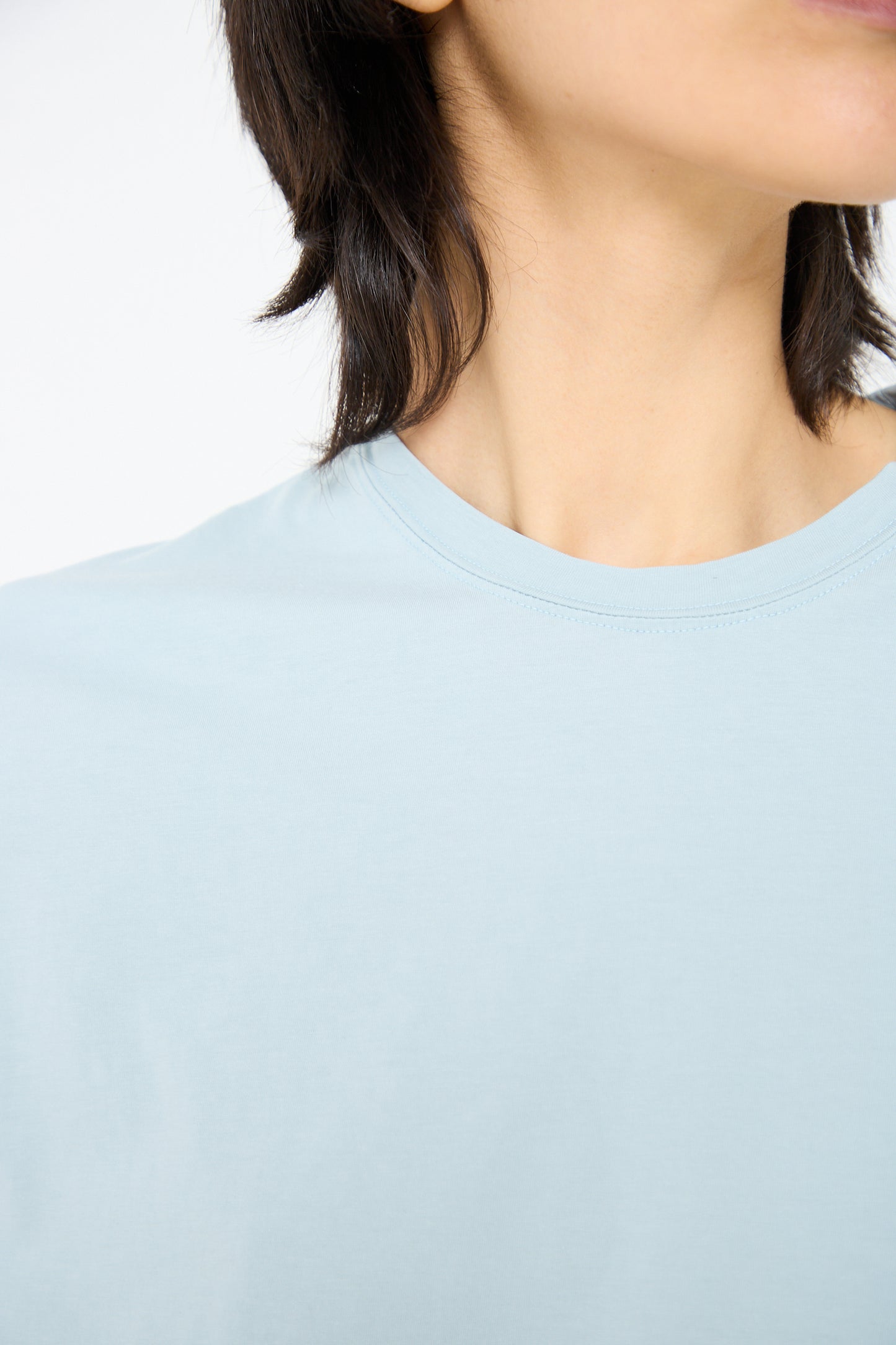 Close-up of a person wearing an Ichi Antiquités Cotton Long Sleeve Pullover Tee in Blue. Only the lower part of their face and neck are visible, with short dark hair falling around their neck. The background is plain white.