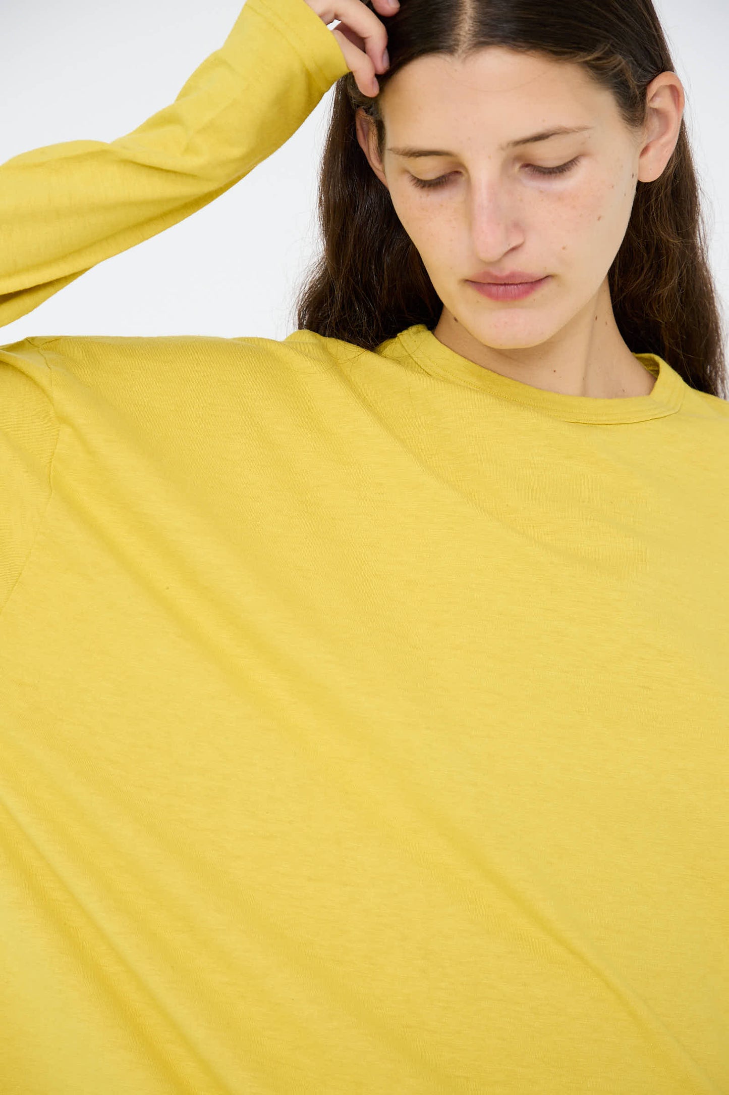 A person wearing the Ichi Antiquités Cotton Loose Long Sleeve Pullover in Mustard looks downward, with one hand raised to their head.