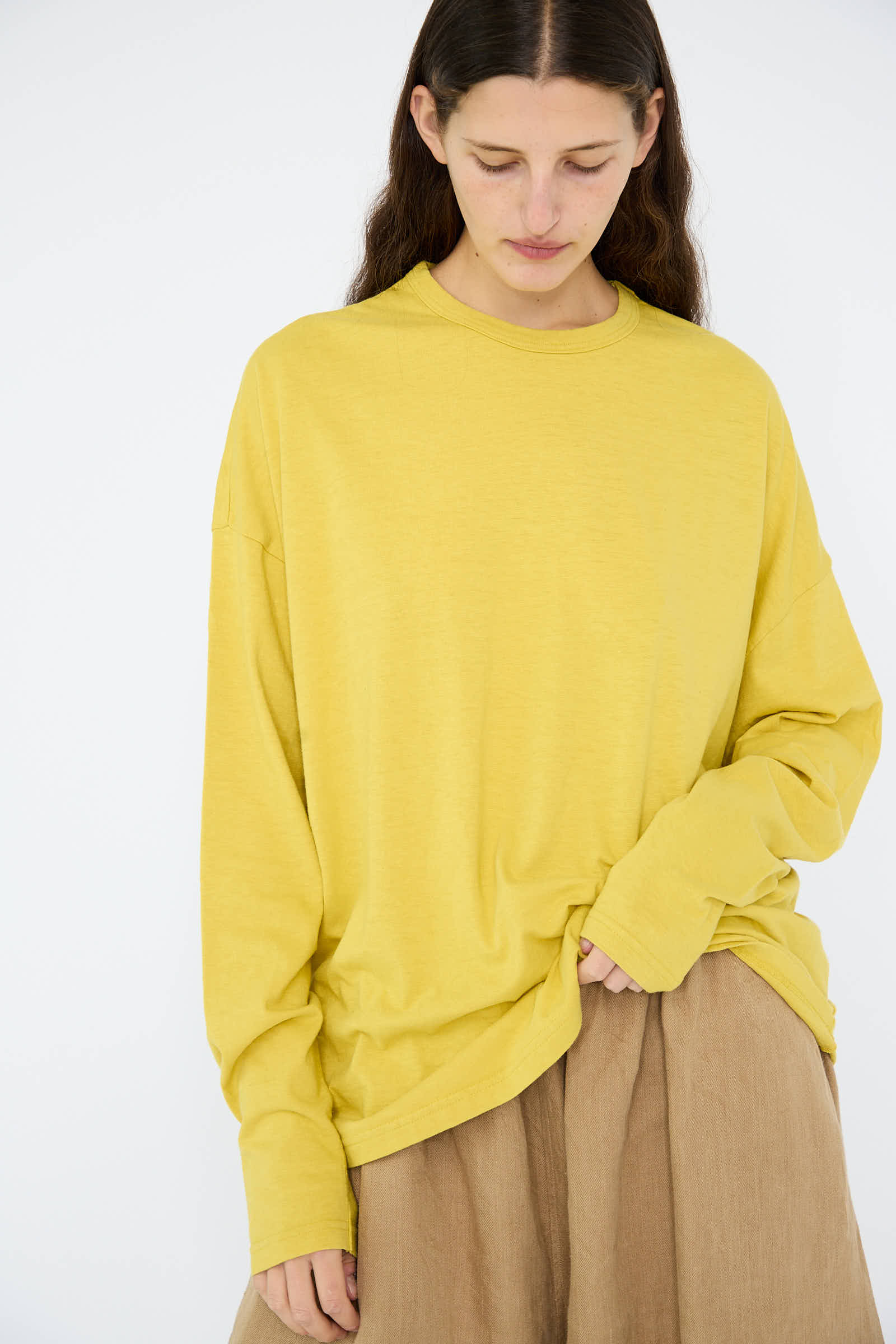 A person with long hair wearing the Ichi Antiquités Cotton Loose Long Sleeve Pullover in Mustard and beige pants stands against a plain background, looking down.