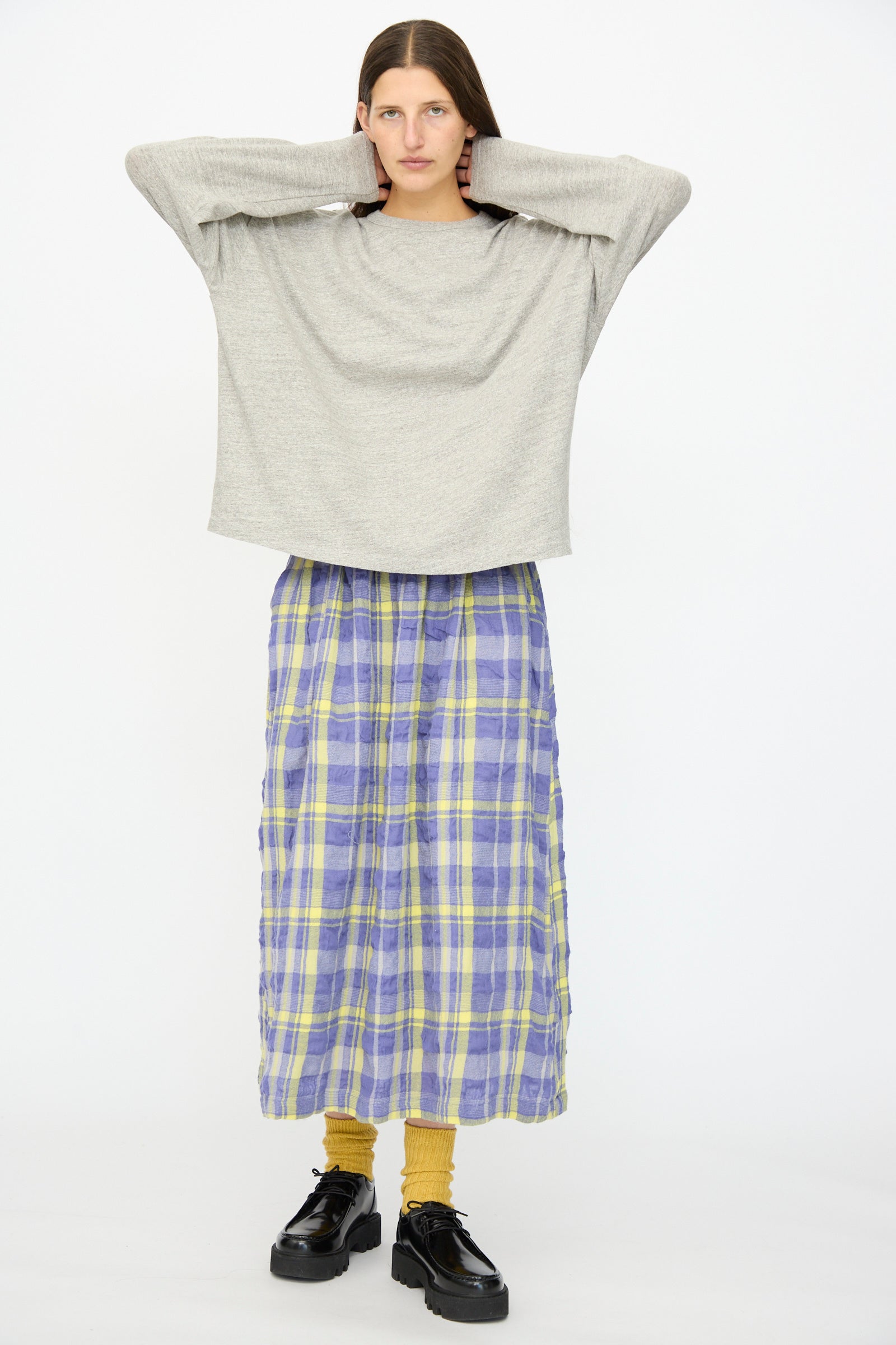 A person stands with hands behind head, wearing the Ichi Antiquités Cotton Loose Pullover in Gray, a long purple and yellow plaid skirt, mustard socks, and black platform shoes against a plain white background.