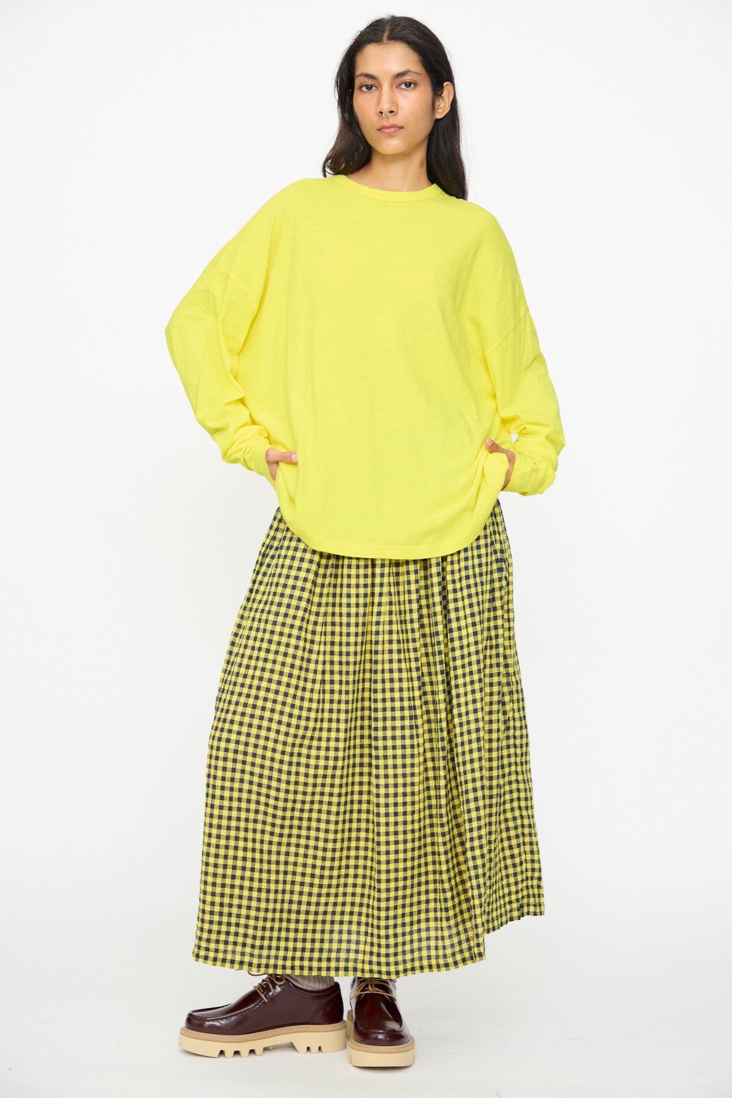 A person stands against a plain white background wearing the Ichi Antiquités Cotton Loose Pullover in Yellow, paired with a yellow and black checkered maxi skirt and brown platform shoes.