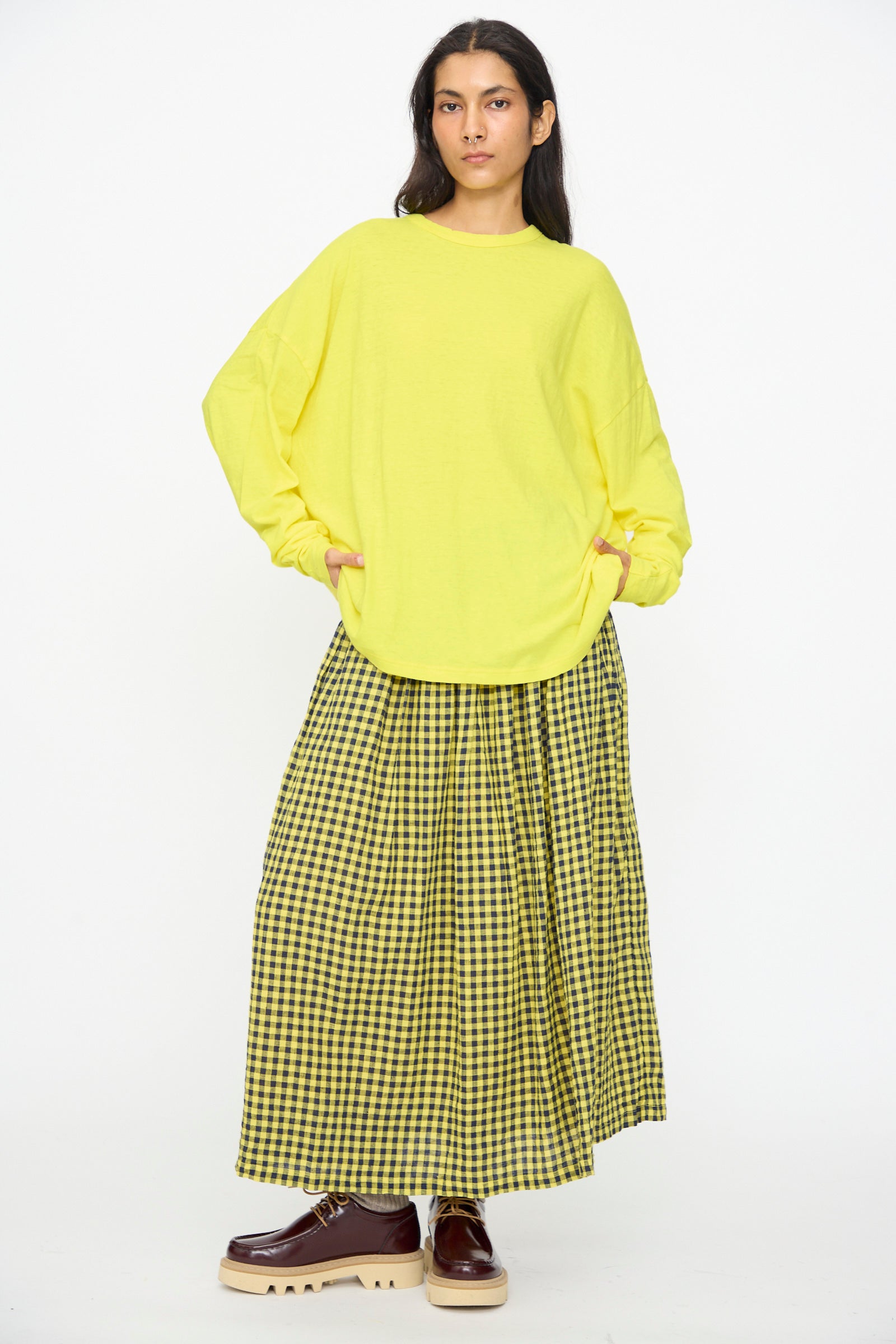 A person stands against a plain white background wearing the Ichi Antiquités Cotton Loose Pullover in Yellow, paired with a yellow and black checkered maxi skirt and brown platform shoes.