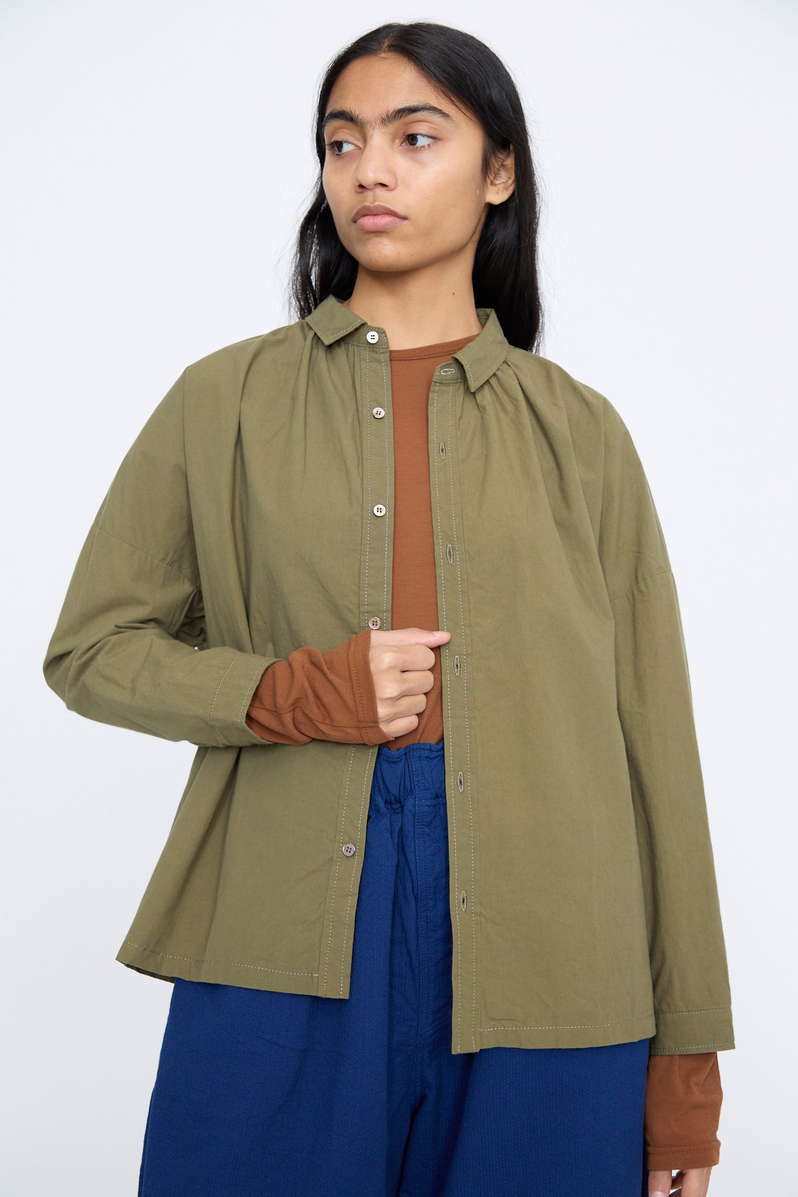 A person in an olive Cotton Ohmi Typewriter Shirt by Ichi Antiquités, layered over a brown top and paired with blue pants, stands against a plain background.