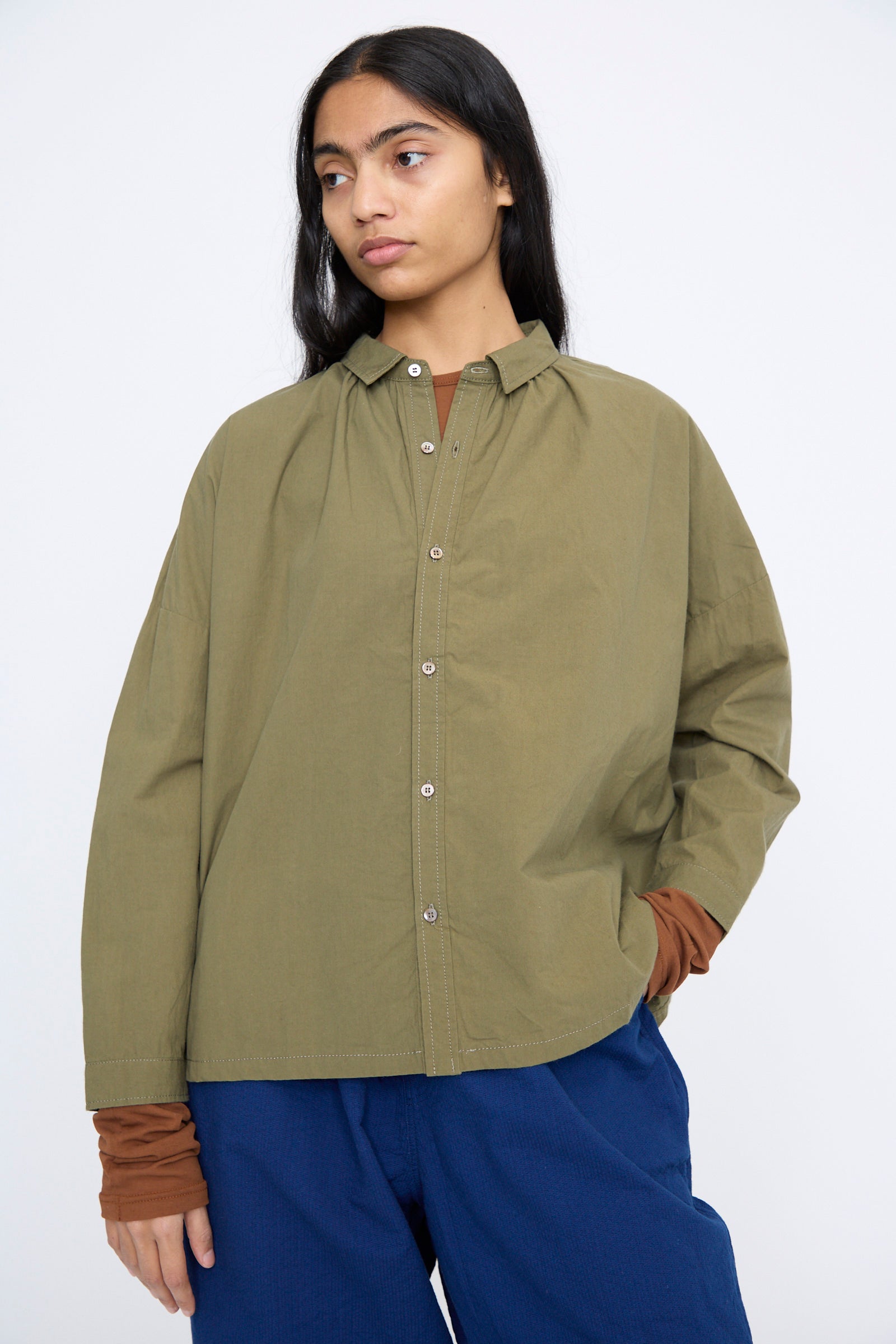 Wearing the Cotton Ohmi Typewriter Shirt in Olive by Ichi Antiquités and blue pants, a person stands against a plain background.
