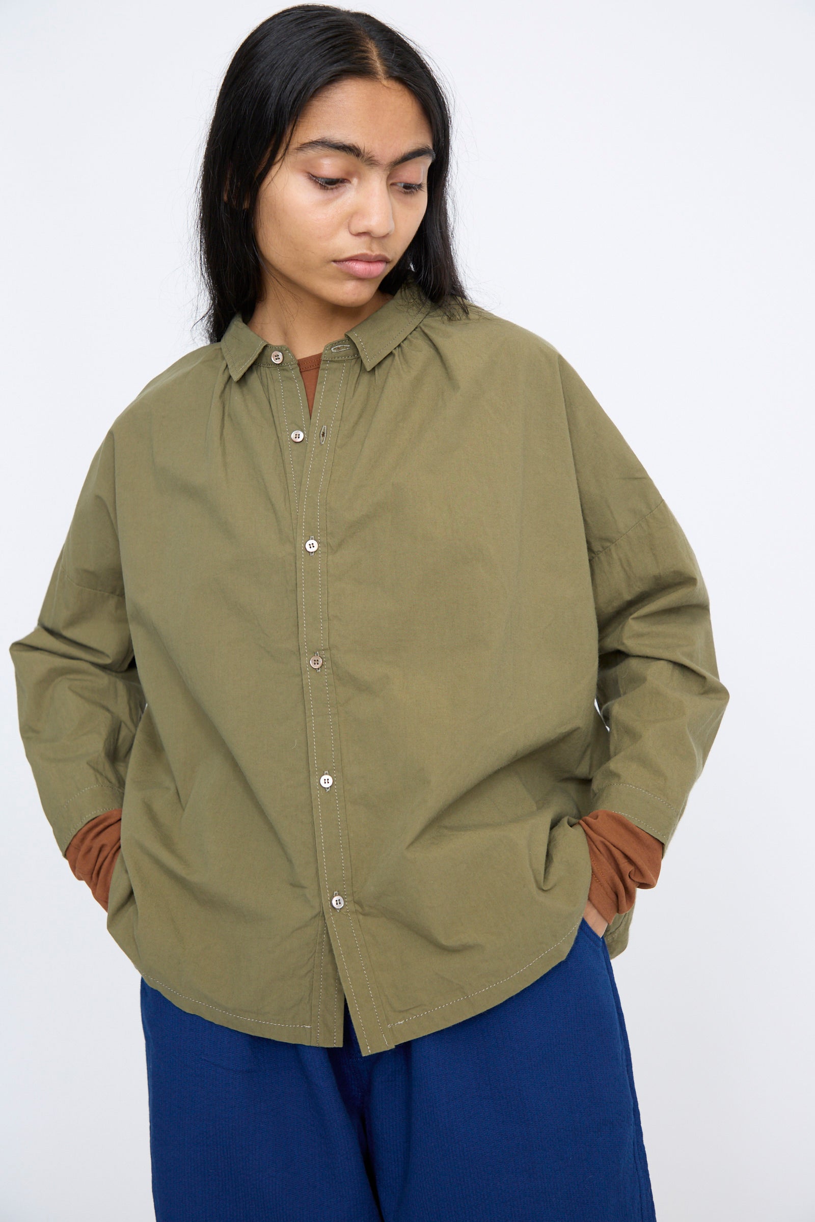 A person in a Cotton Ohmi Typewriter Shirt by Ichi Antiquités, olive, relaxed fit with long sleeves, hands in pockets, looking down against a plain background.
