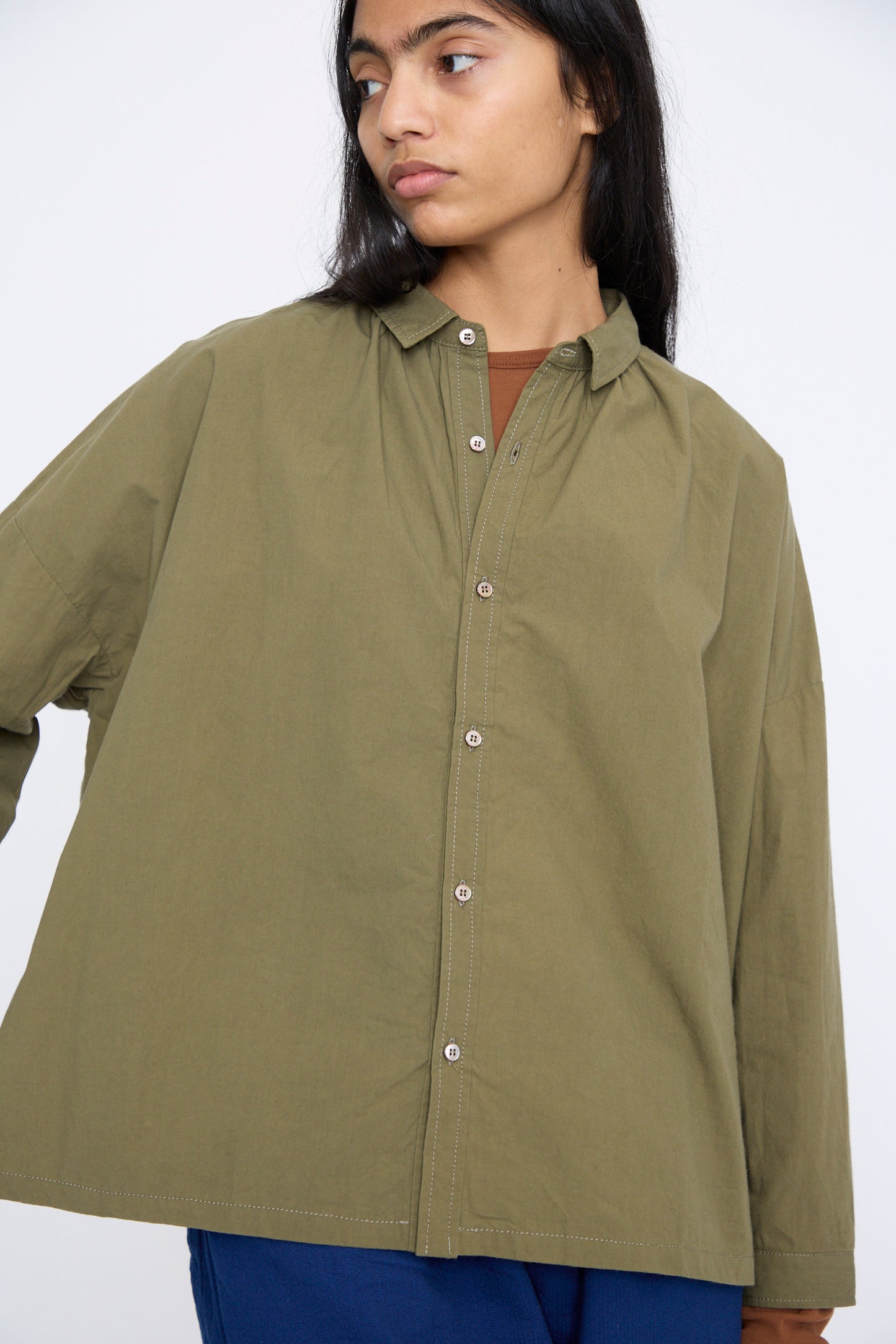 Wearing a relaxed fit Ichi Antiquités Cotton Ohmi Typewriter Shirt in olive and blue pants, the individual stands against a plain background, gazing to the side.