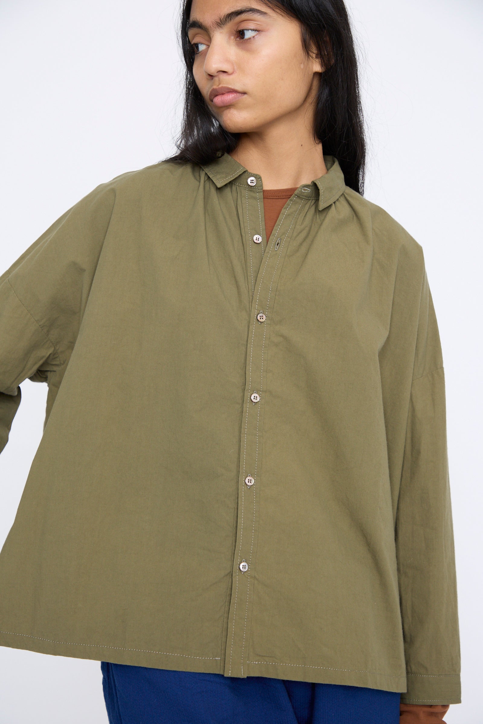 Wearing a relaxed fit Ichi Antiquités Cotton Ohmi Typewriter Shirt in olive and blue pants, the individual stands against a plain background, gazing to the side.