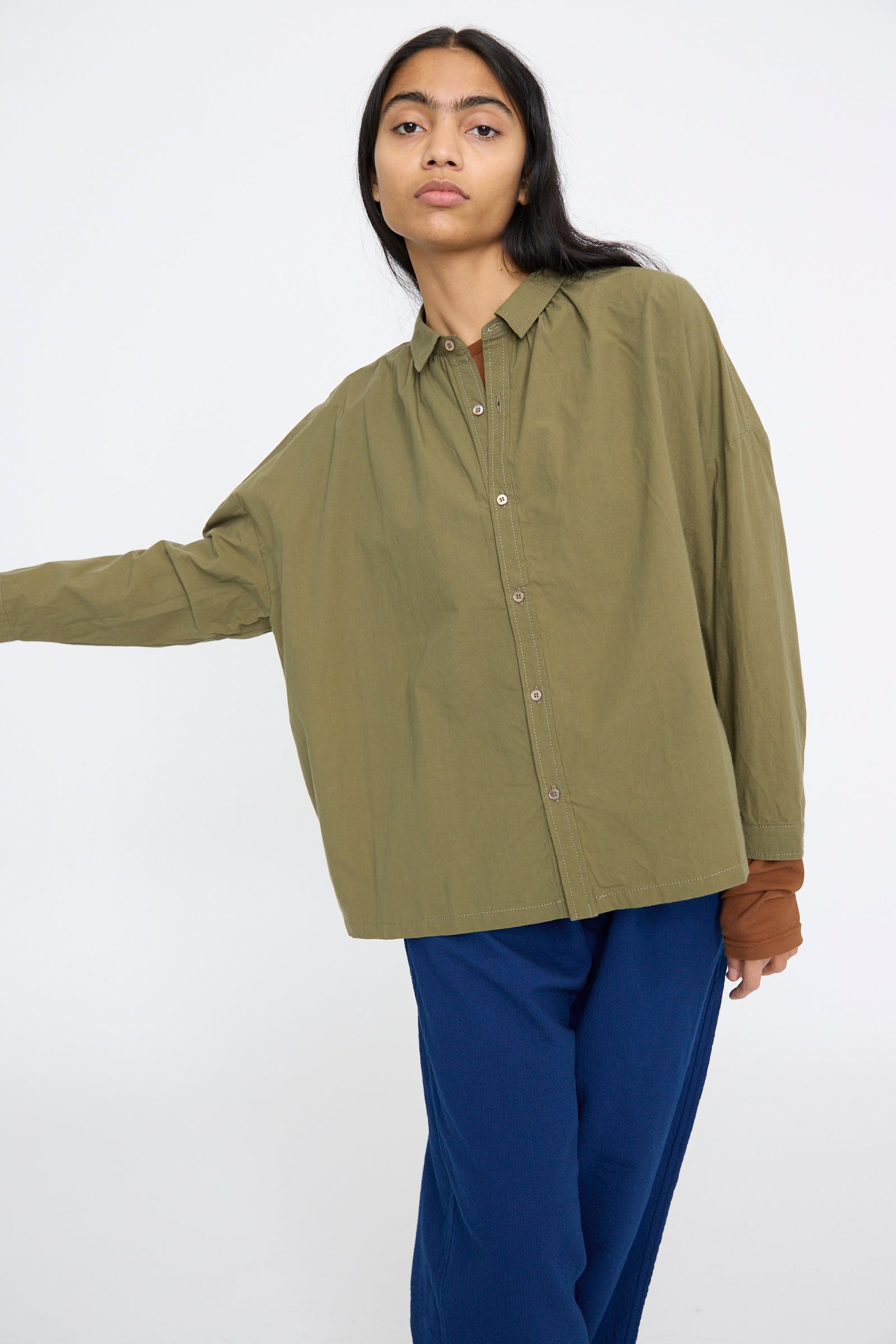 Dressed in an oversized Cotton Ohmi Typewriter Shirt in Olive by Ichi Antiquités and relaxed fit blue pants, the person stands forward against a white backdrop.