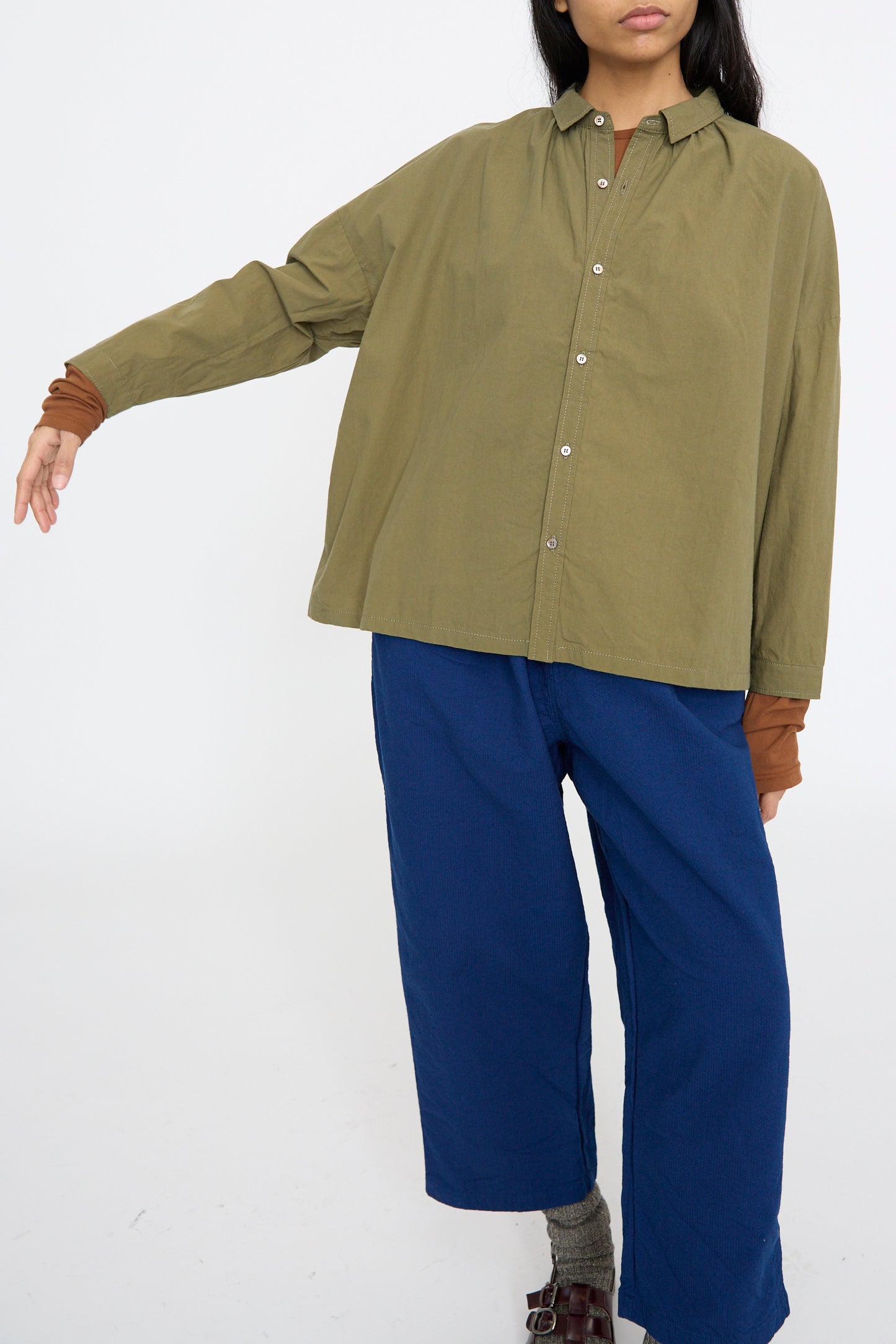 A person wearing the Ichi Antiquités Cotton Ohmi Typewriter Shirt in Olive and blue pants stands against a plain white background with one arm extended.