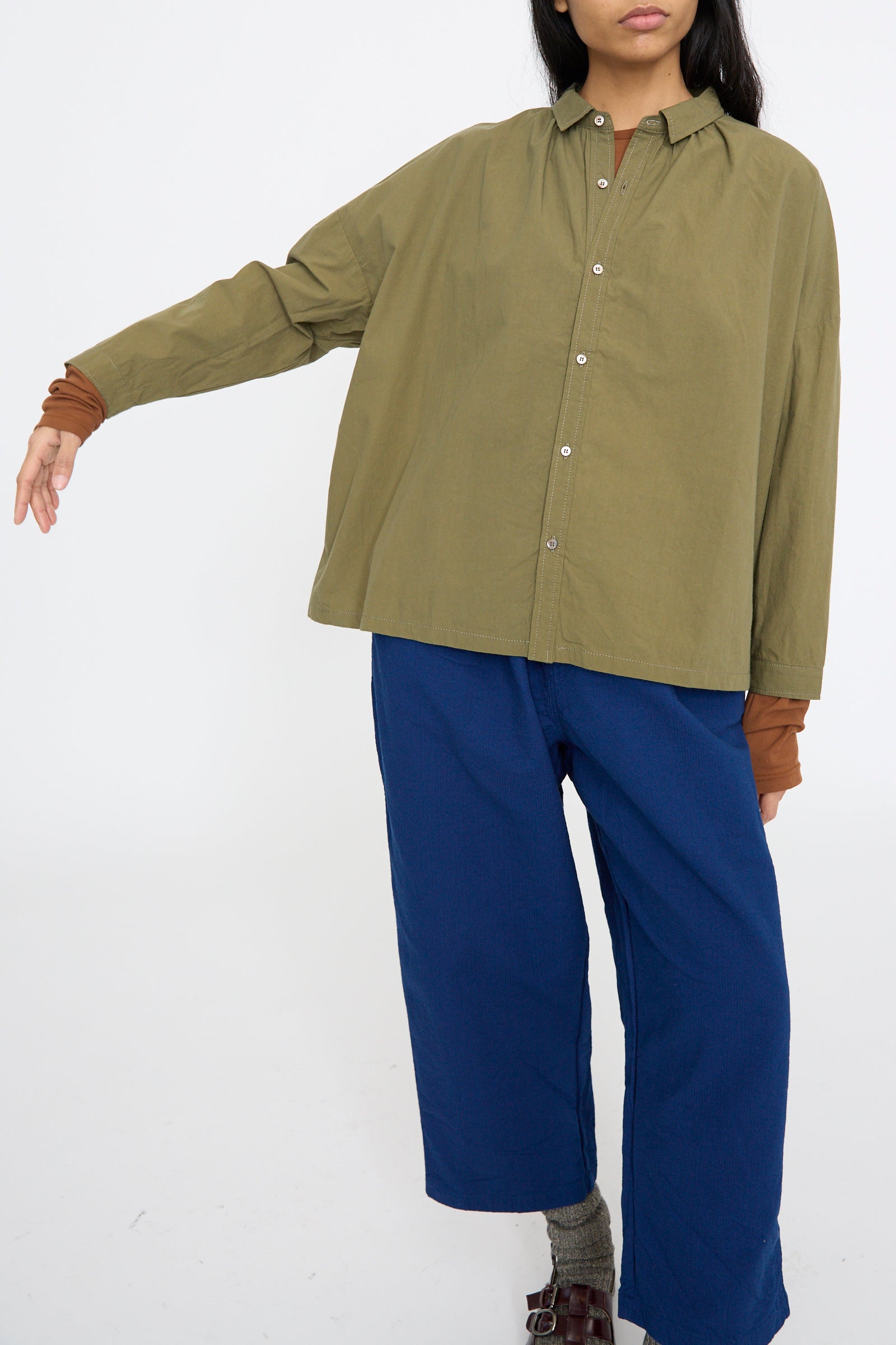 A person wearing the Ichi Antiquités Cotton Ohmi Typewriter Shirt in Olive and blue pants stands against a plain white background with one arm extended.