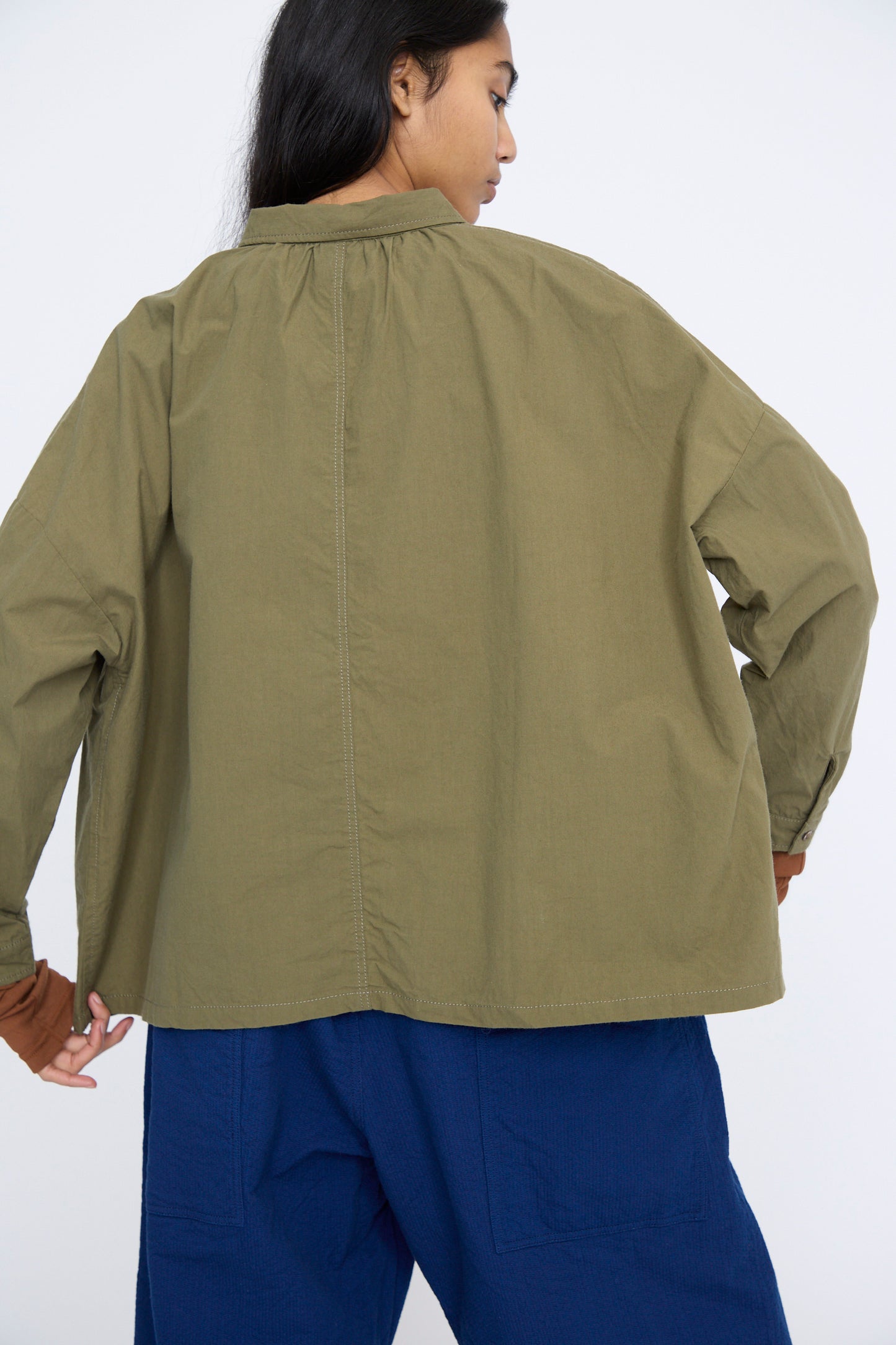 Facing away, a person with long dark hair wears the Ichi Antiquités Cotton Ohmi Typewriter Shirt in Olive paired with blue pants against a white background.
