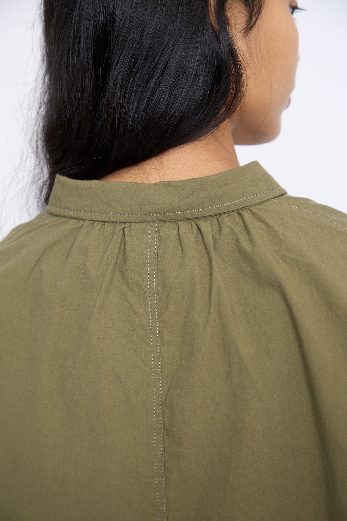 Close-up of a person with long black hair wearing the Cotton Ohmi Typewriter Shirt in Olive by Ichi Antiquités, highlighting its relaxed fit collar and intricate stitching on the long sleeve cotton garment.