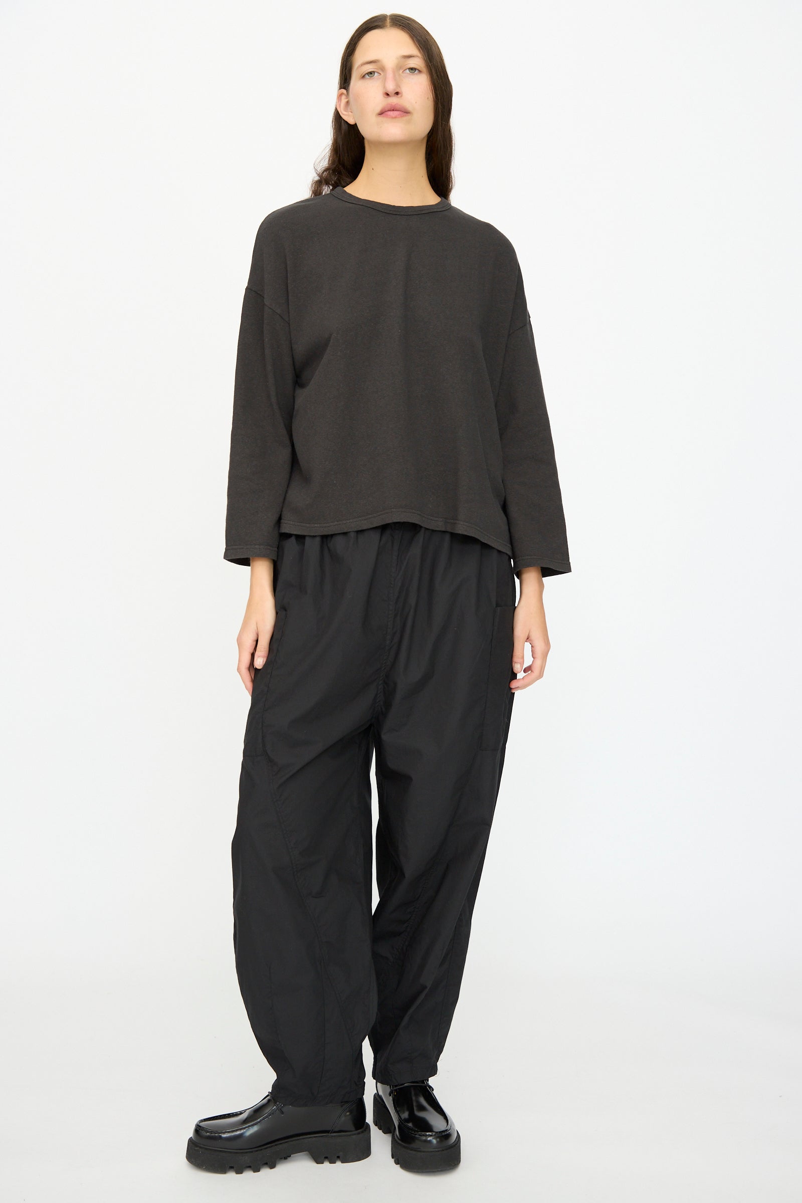 A person stands against a plain white background wearing the Ichi Antiquités Cotton Pullover in Black, paired with baggy black pants and black platform shoes.