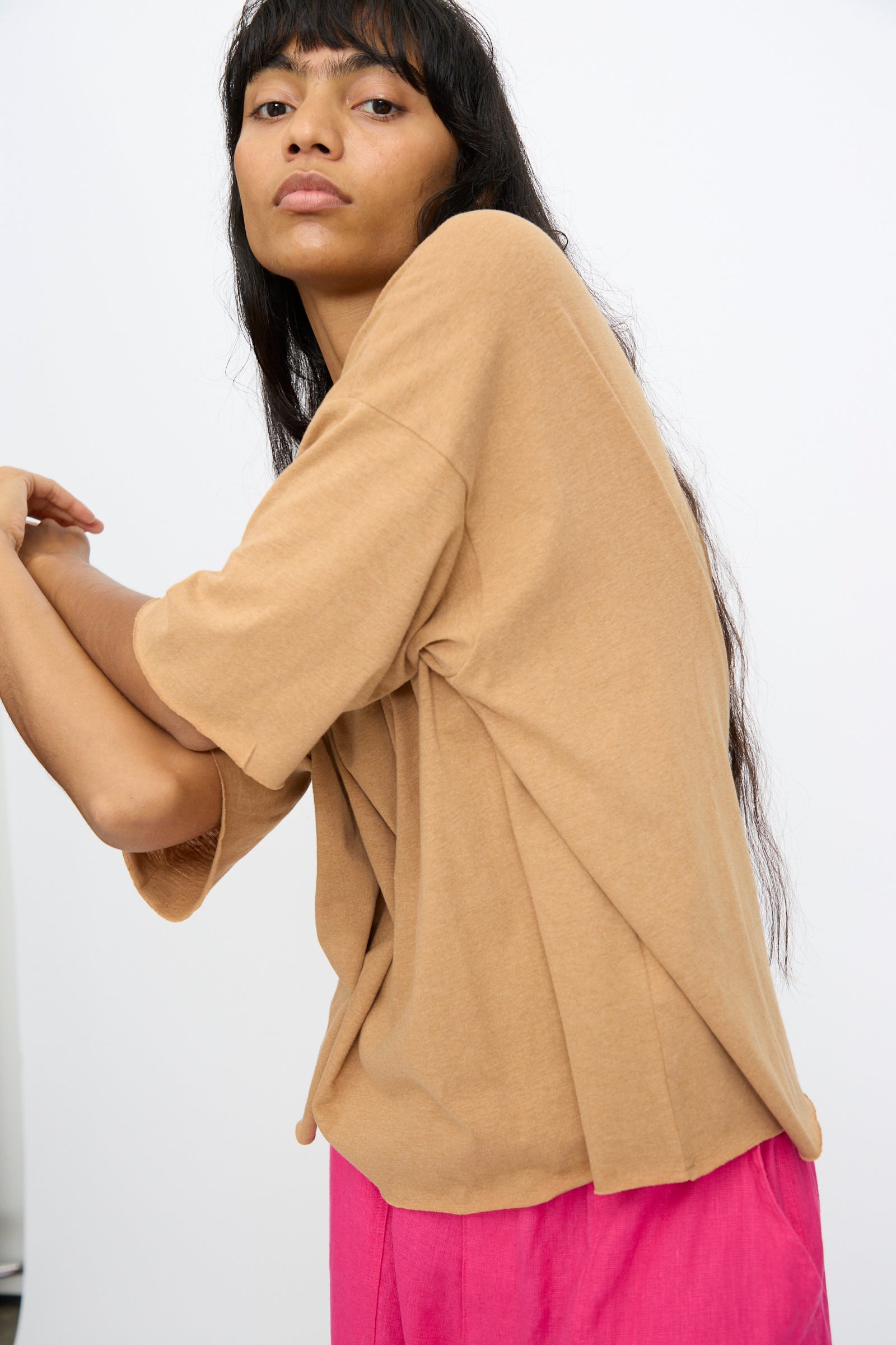 A woman with long dark hair models the Ichi Antiquités Cotton Pullover Tee in Camel, styled with bright pink pants, posing against a plain white background.