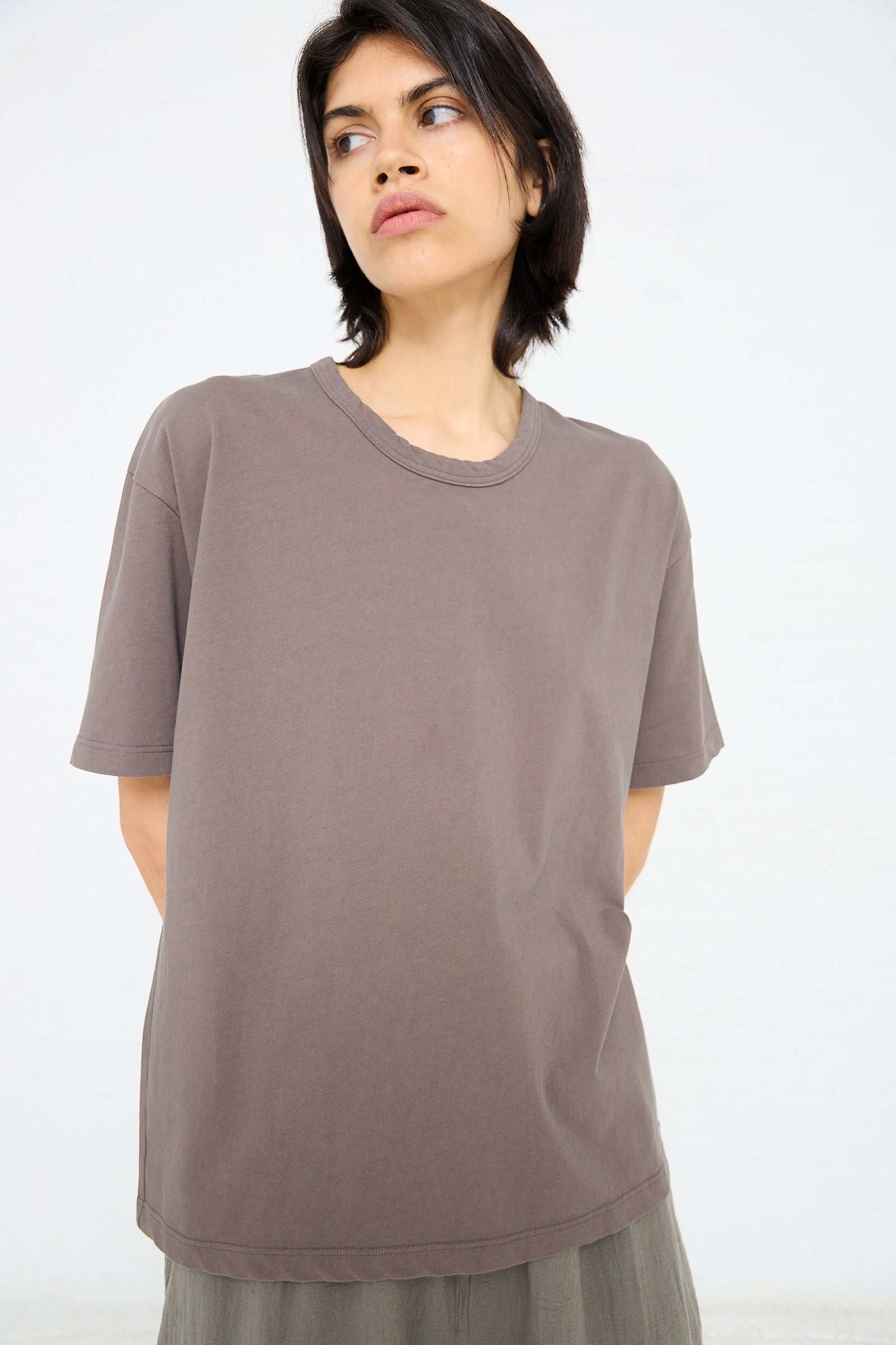 A person with short dark hair poses against a white background, wearing a loose-fitting Cotton Pullover Tee in Mocha from Ichi Antiquités with a wide crew neck and light-colored pants.