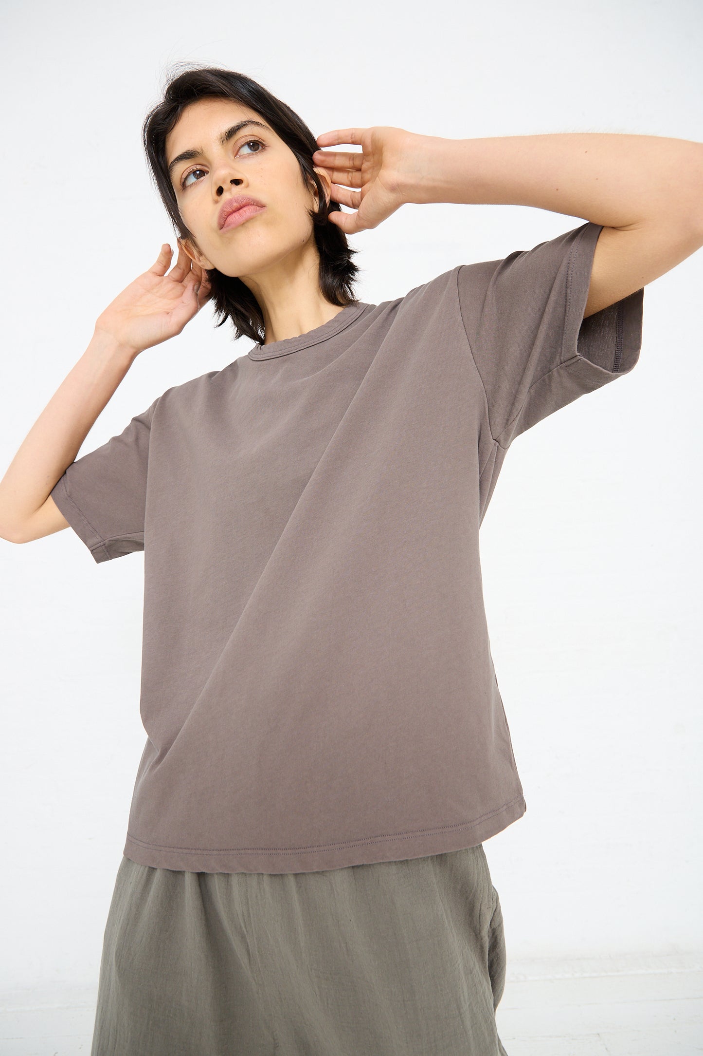 Person wearing a loose-fitting, Ichi Antiquités Cotton Pullover Tee in Mocha and gray pants, with their arms bent and hands positioned near their neck. The background is plain white.