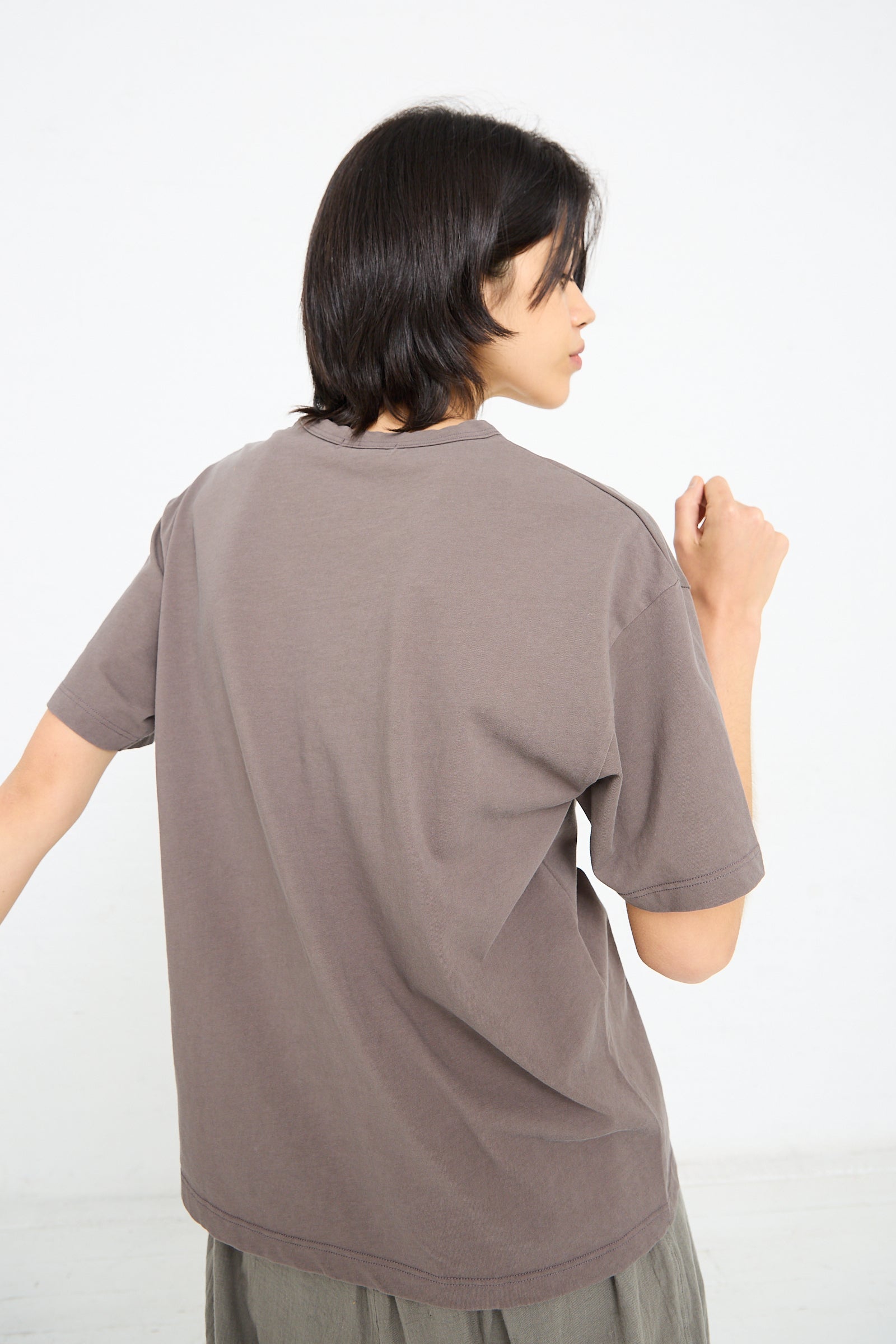 A person with medium-length mocha brown hair is standing with their back facing the camera, wearing a plain grey **Cotton Pullover Tee in Mocha by Ichi Antiquités** and dark-colored pants.