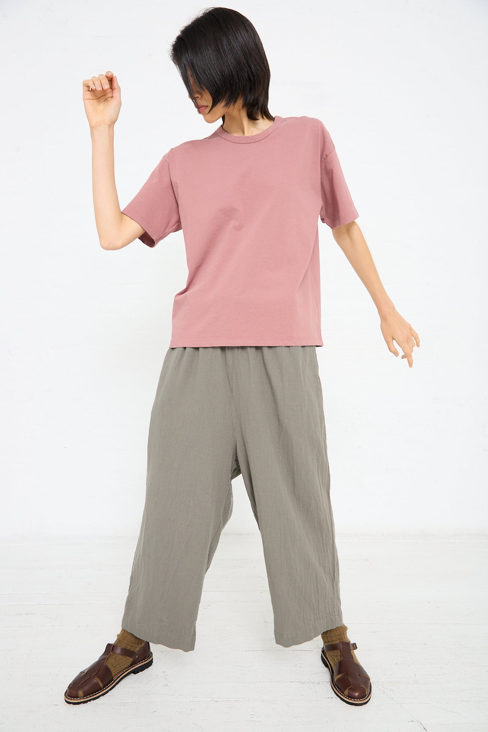 A person stands against a white background, wearing an Ichi Antiquités Cotton Pullover Tee in Pink, loose gray pants, and brown shoes. They have their right hand raised and appear to be in mid-movement.