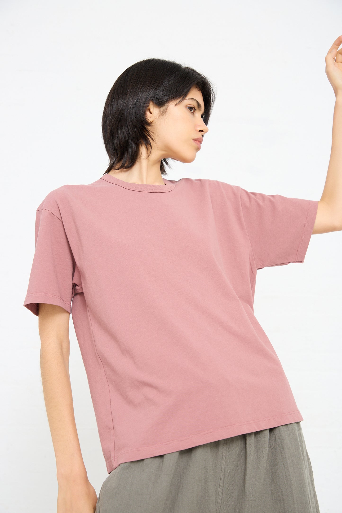 A person with short dark hair wears a loose-fitting pink t-shirt, posing with their left arm raised against a plain white background. The Ichi Antiquités Cotton Pullover Tee in Pink boasts a comfortable boxy fit.