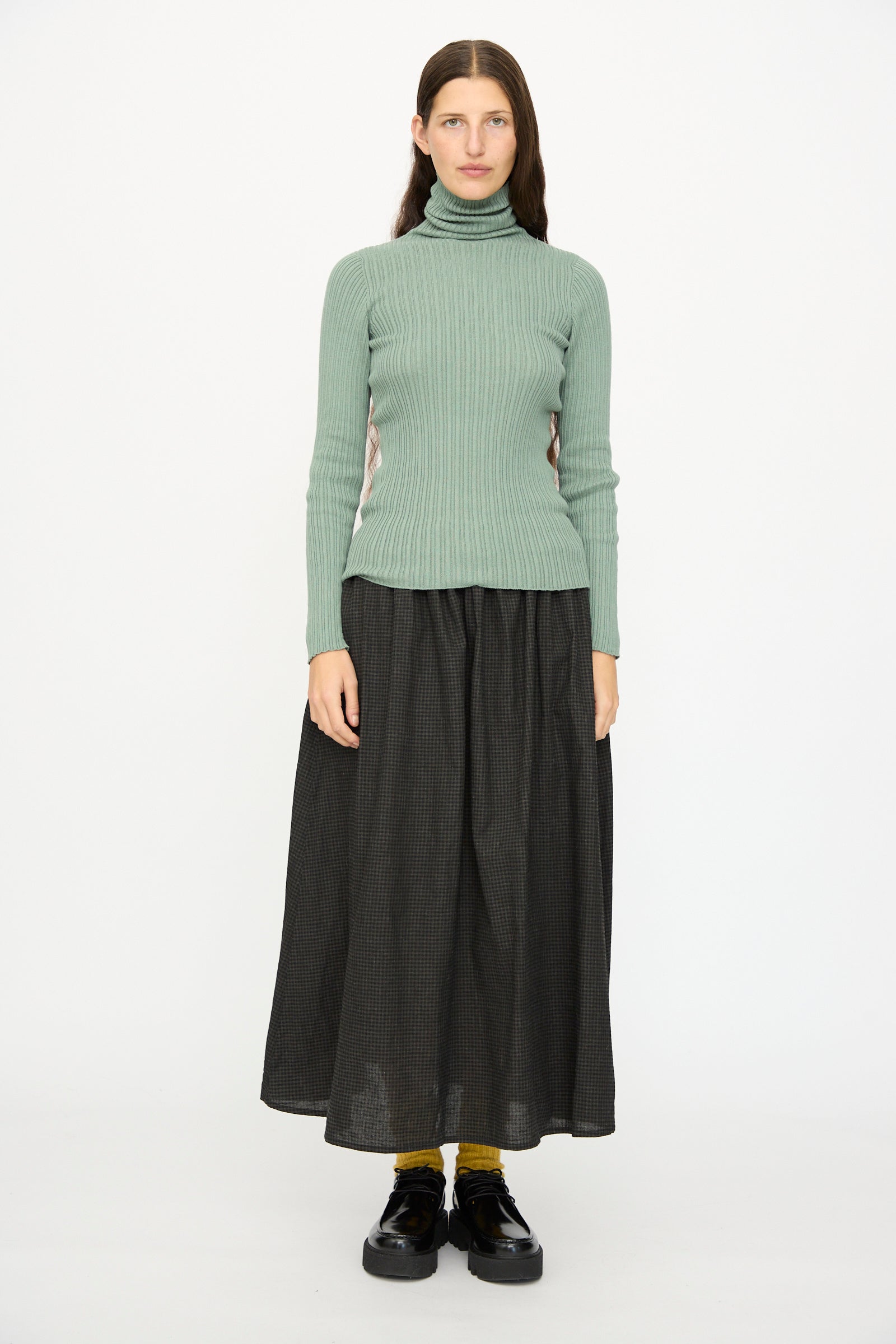 A person faces forward against a white background, wearing the Ichi Antiquités Cotton Rib Knit Turtleneck in Green, a long dark skirt, yellow socks, and black shoes.