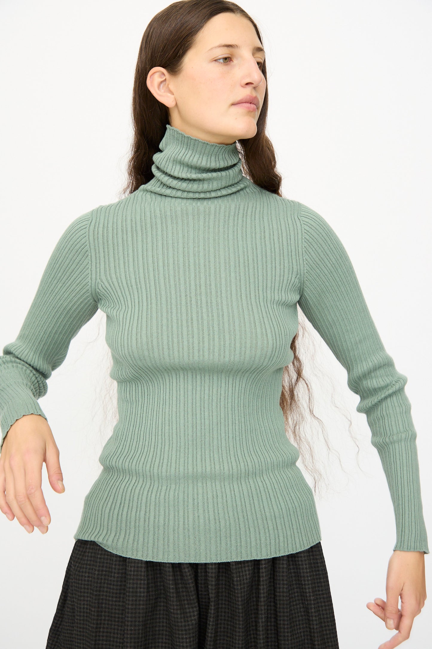 A woman with long brown hair wears the Ichi Antiquités Cotton Rib Knit Turtleneck in Green, styled with a dark skirt and standing against a plain light background.