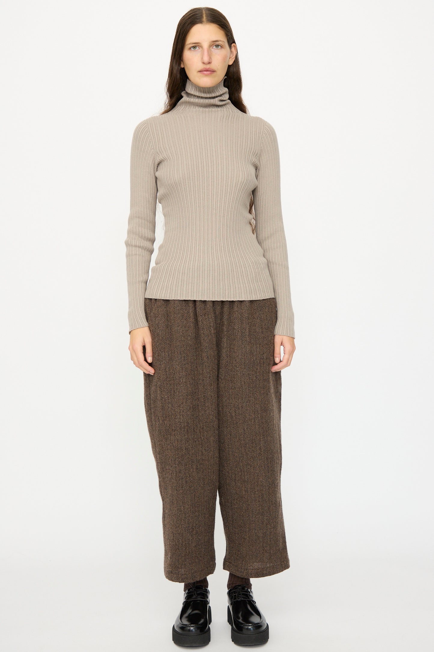 A woman faces forward against a white background, wearing the Ichi Antiquités Cotton Rib Knit Turtleneck in Mocha, loose brown pants, and black shoes.
